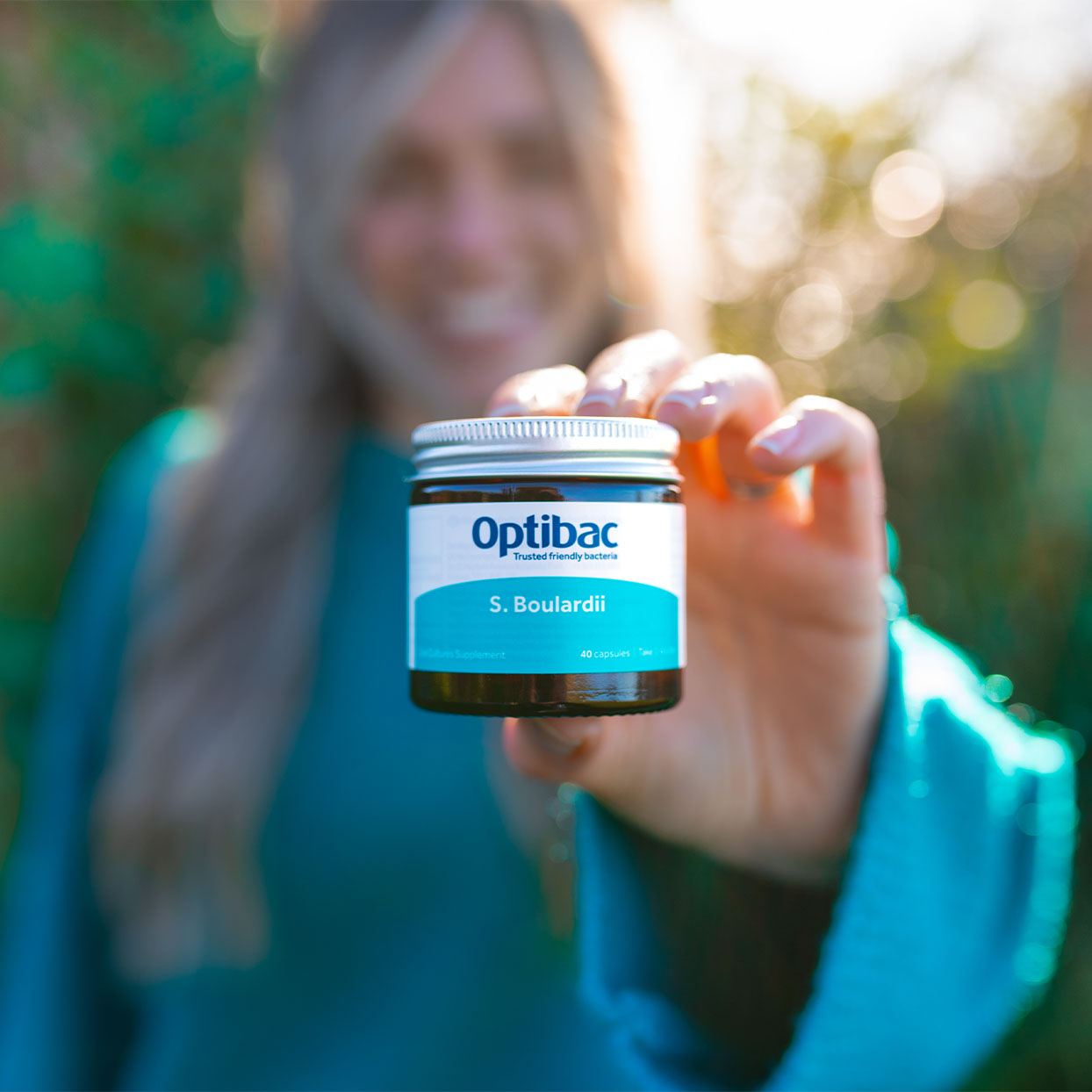 Person holding a jar of Optibac S. Boulardii supplements outdoors, smiling. The label is clearly visible.