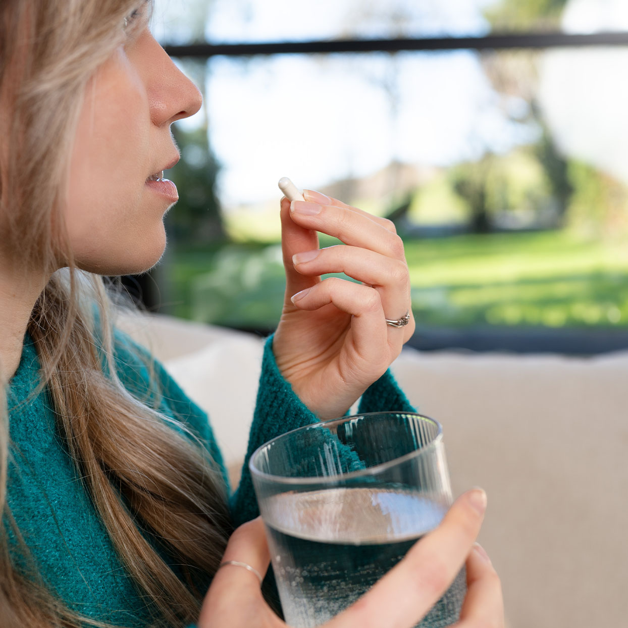 A person in a green sweater holds a pill near their mouth and a glass of water, sitting indoors with a blurred outdoor view in the background.