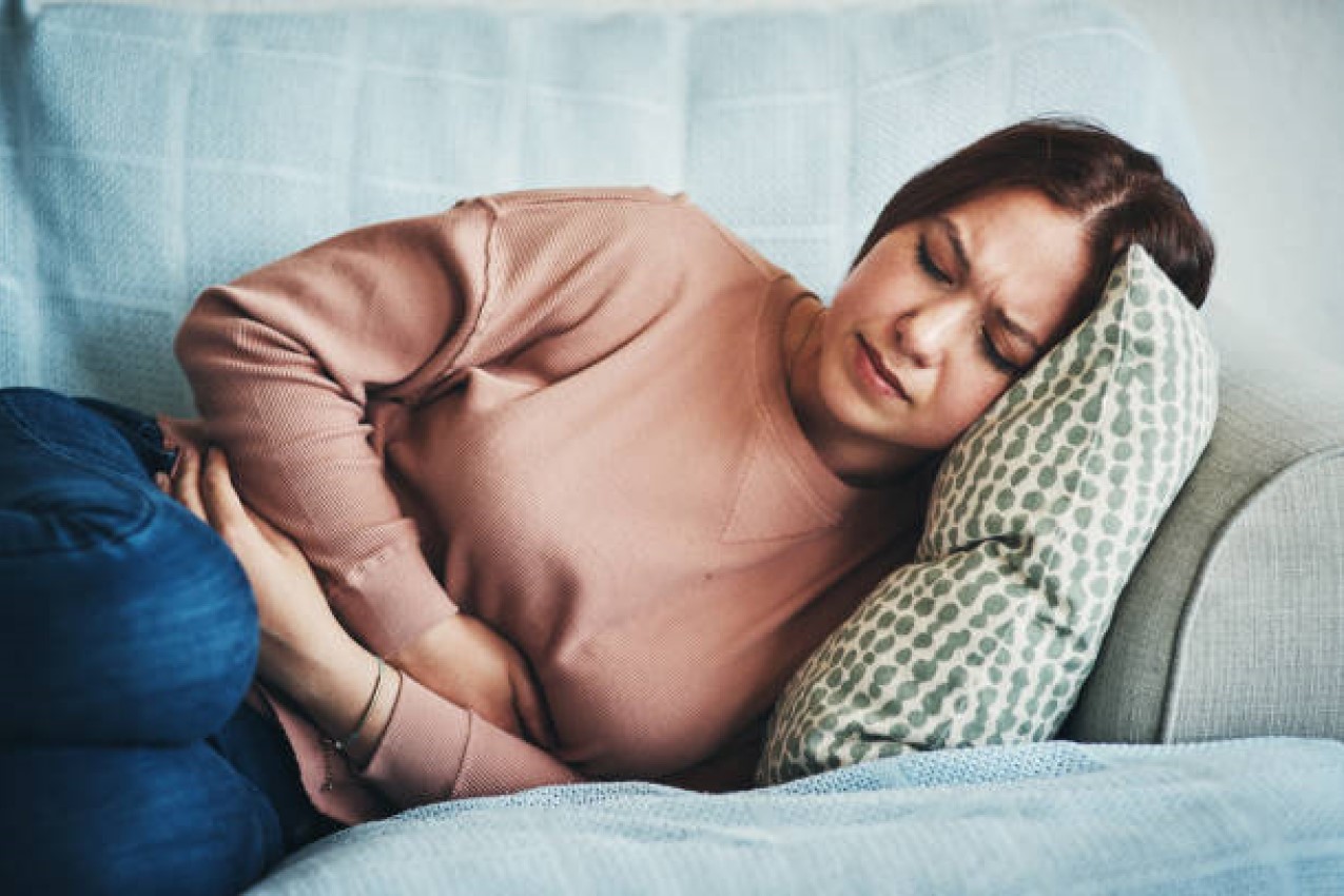Woman lying on a couch, holding her stomach in discomfort, wearing a pink sweater and blue jeans, with a patterned pillow under her head.
