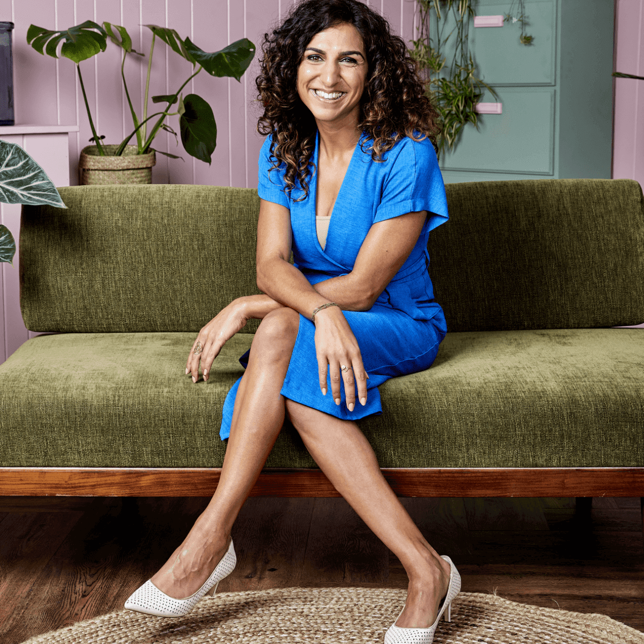 Smiling woman in a blue dress sits on a green sofa with plants and pink walls in the background.
