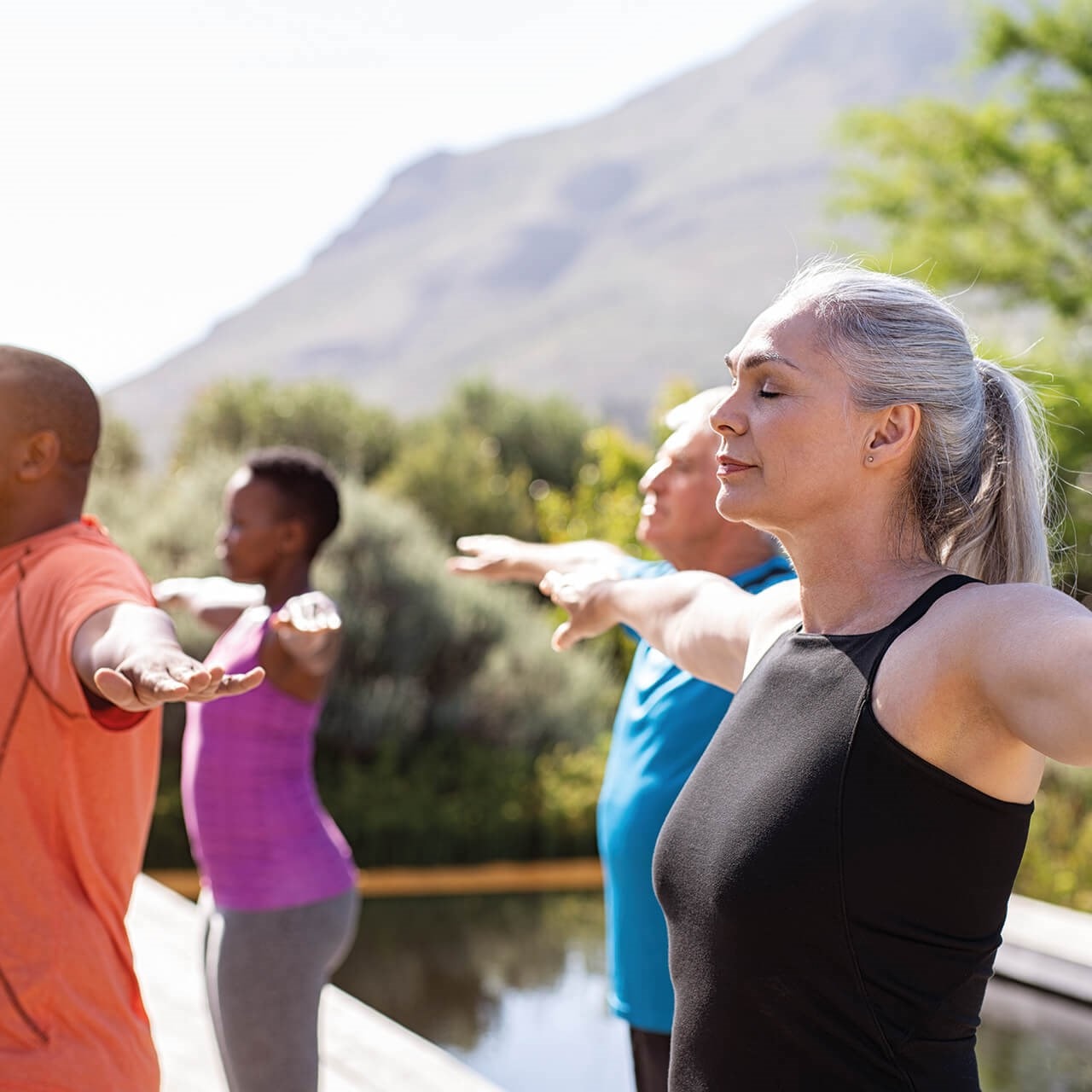 A diverse group of people practicing yoga outdoors, standing with arms extended, eyes closed, in a serene natural setting.