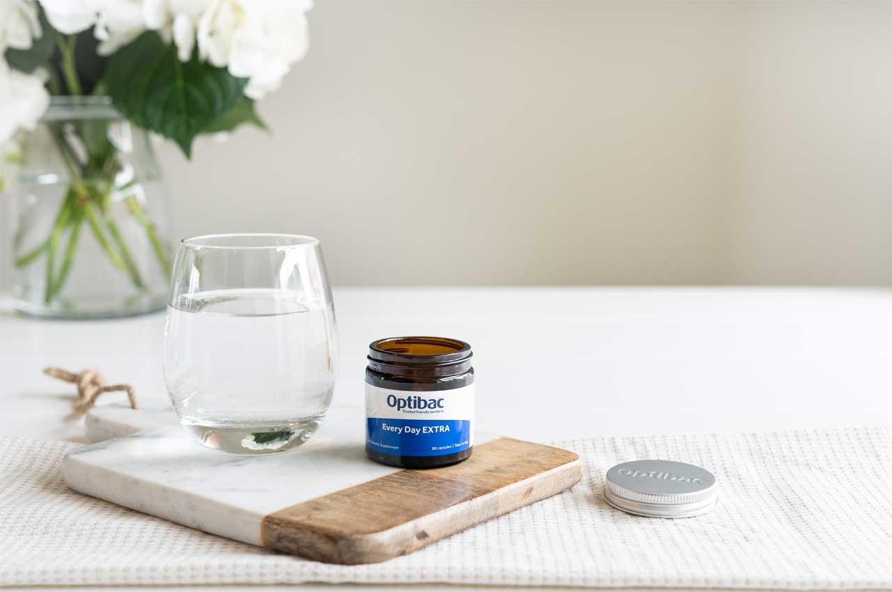 A glass of water and a jar of Optibac supplements on a wooden board, with flowers in the background.