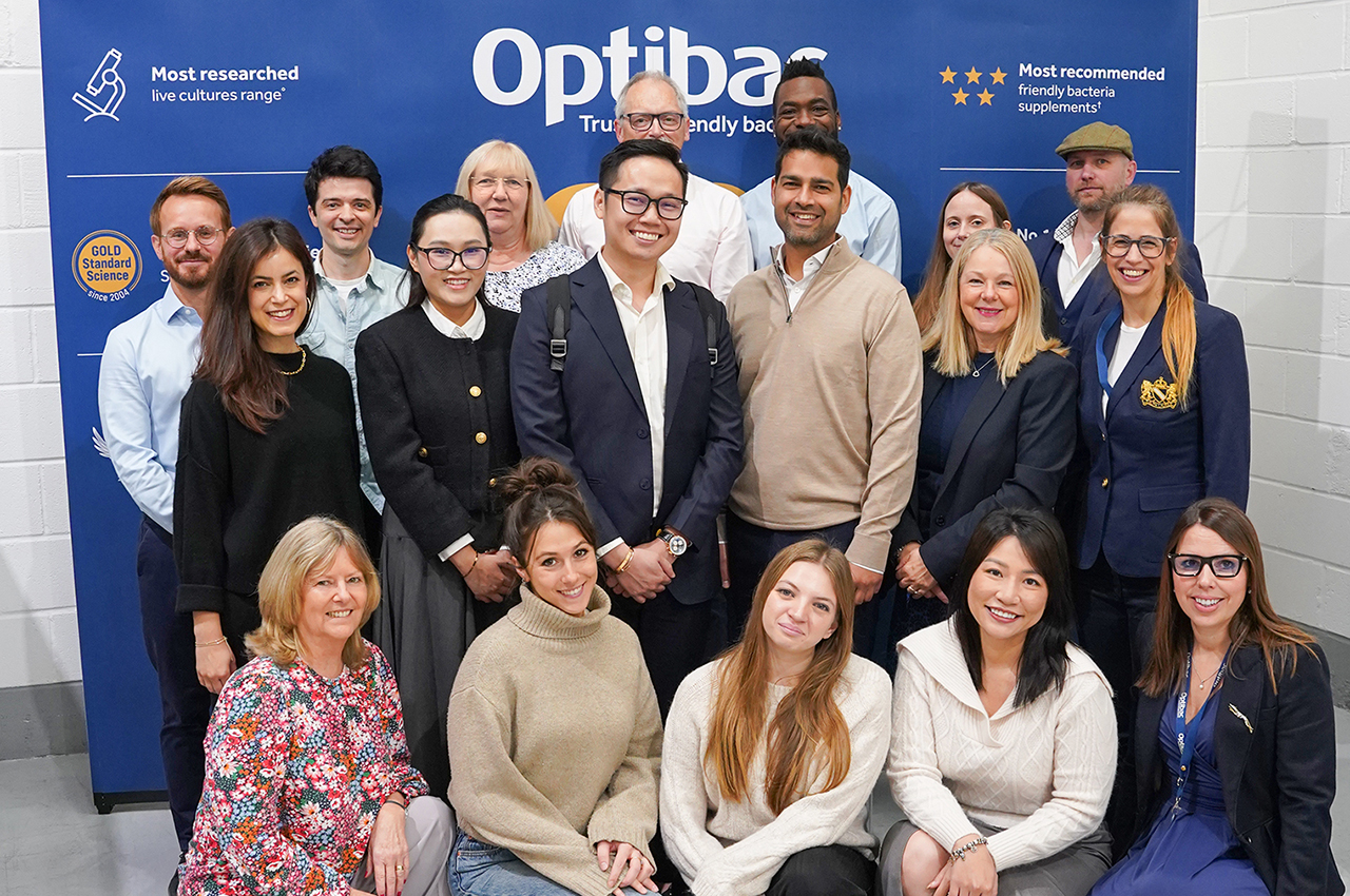 A diverse group of smiling people posing in front of an "Optibac" backdrop with awards and recommendations displayed.
