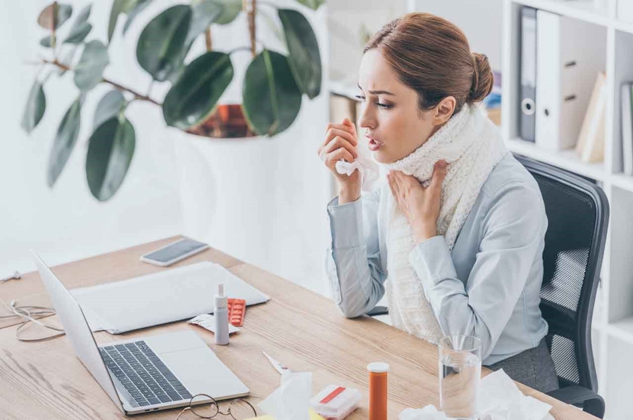 Woman with a scarf around her neck, sitting at a desk with a laptop, tissues, and medicine, appearing unwell.