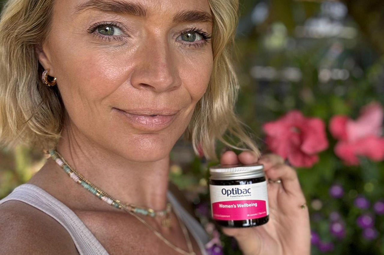 Woman holding a jar of Optibac Women's Wellbeing supplements, standing outdoors with pink flowers in the background.