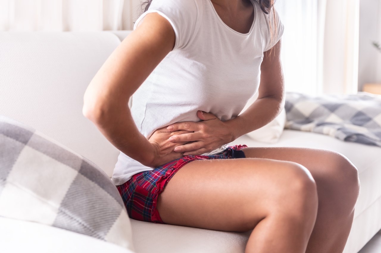 Person sitting on a couch, holding their abdomen in discomfort, wearing a white shirt and plaid shorts.