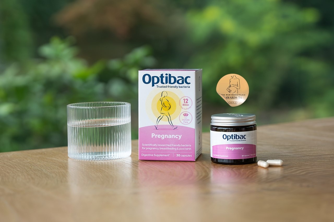 Optibac Pregnancy probiotics box and jar on a wooden table with a glass of water and capsules, set against a blurred green background.