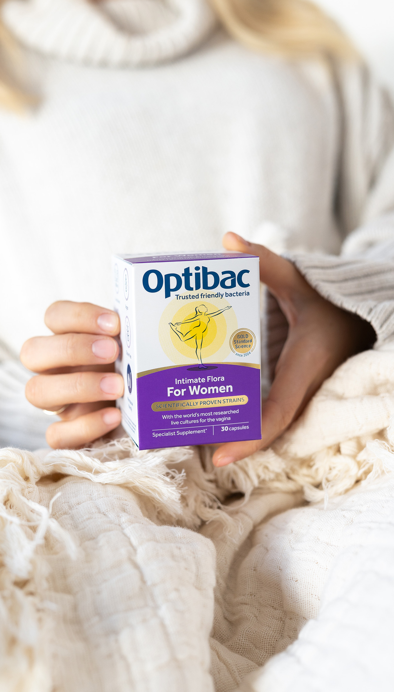 A person in a cozy sweater holds a box of Optibac Intimate Flora for Women, a probiotic supplement.