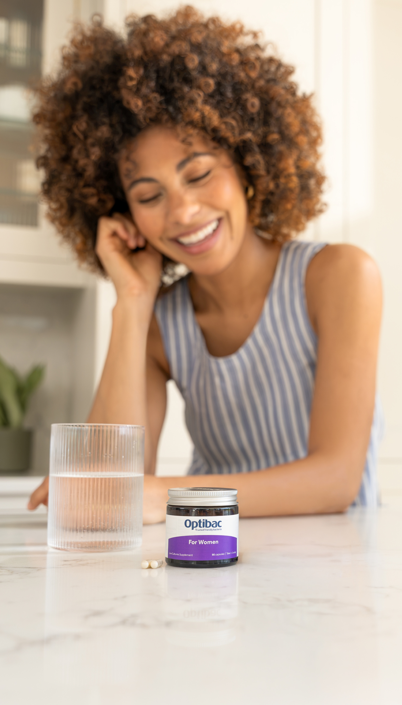 A woman in a striped dress smiles while leaning on a table with a glass of water and a jar of Optibac supplements for women in the foreground.