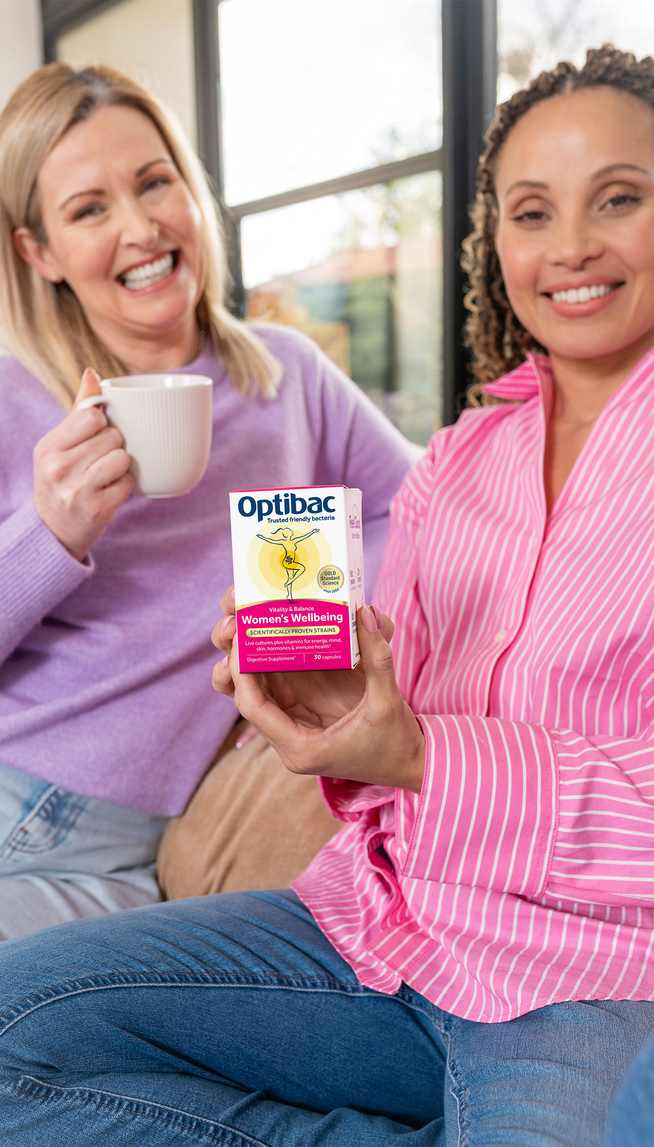 Two women smiling, one holding a box of Optibac Women's Wellbeing supplements, sitting on a couch with a mug.