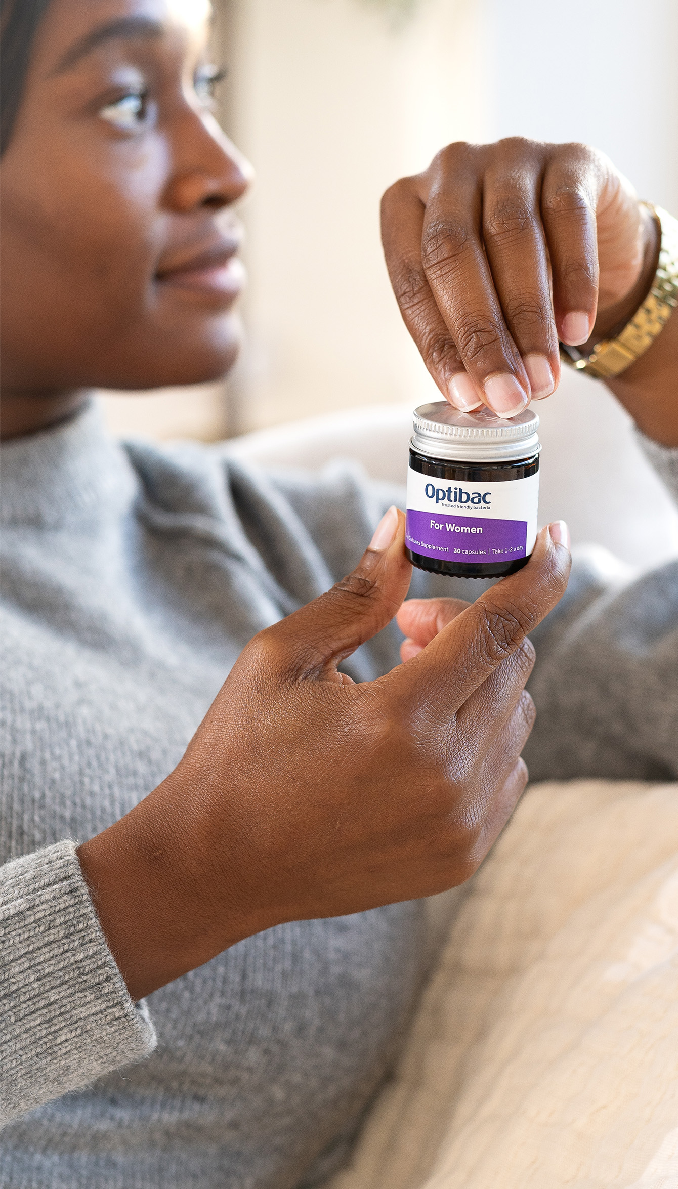 Person holding a small jar labeled "Optibac for Women" with a purple label, wearing a gray sweater and gold watch.