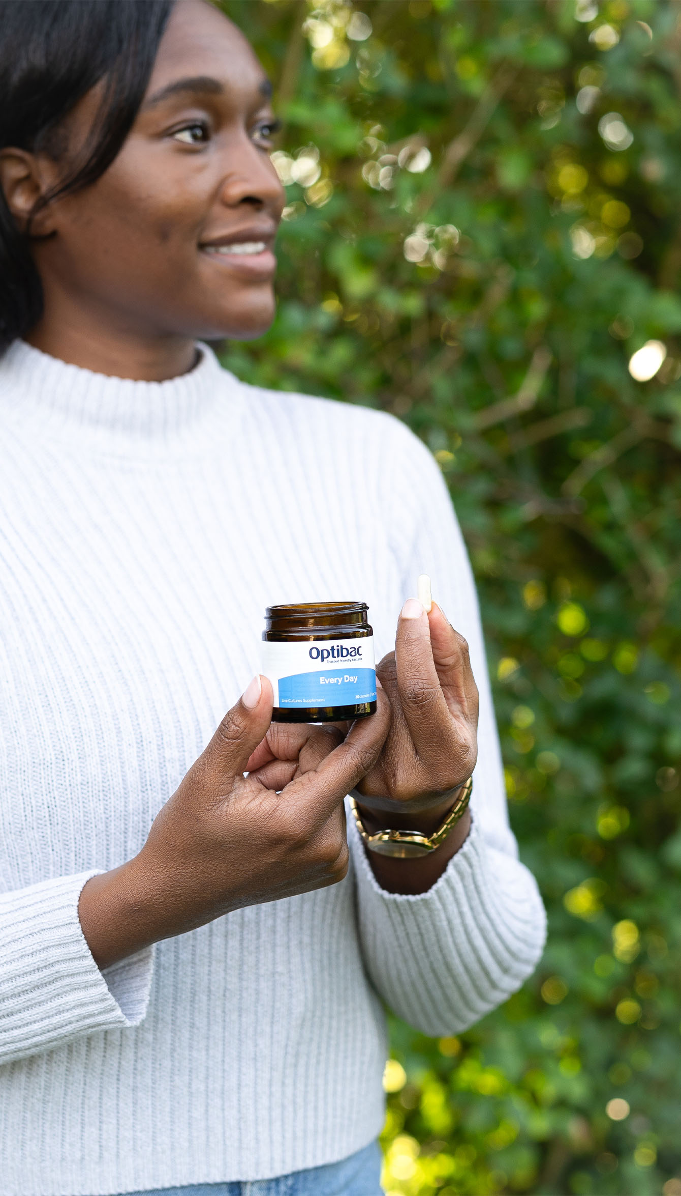 Person holding a jar of Optibac supplements outdoors, wearing a white sweater, with greenery in the background.