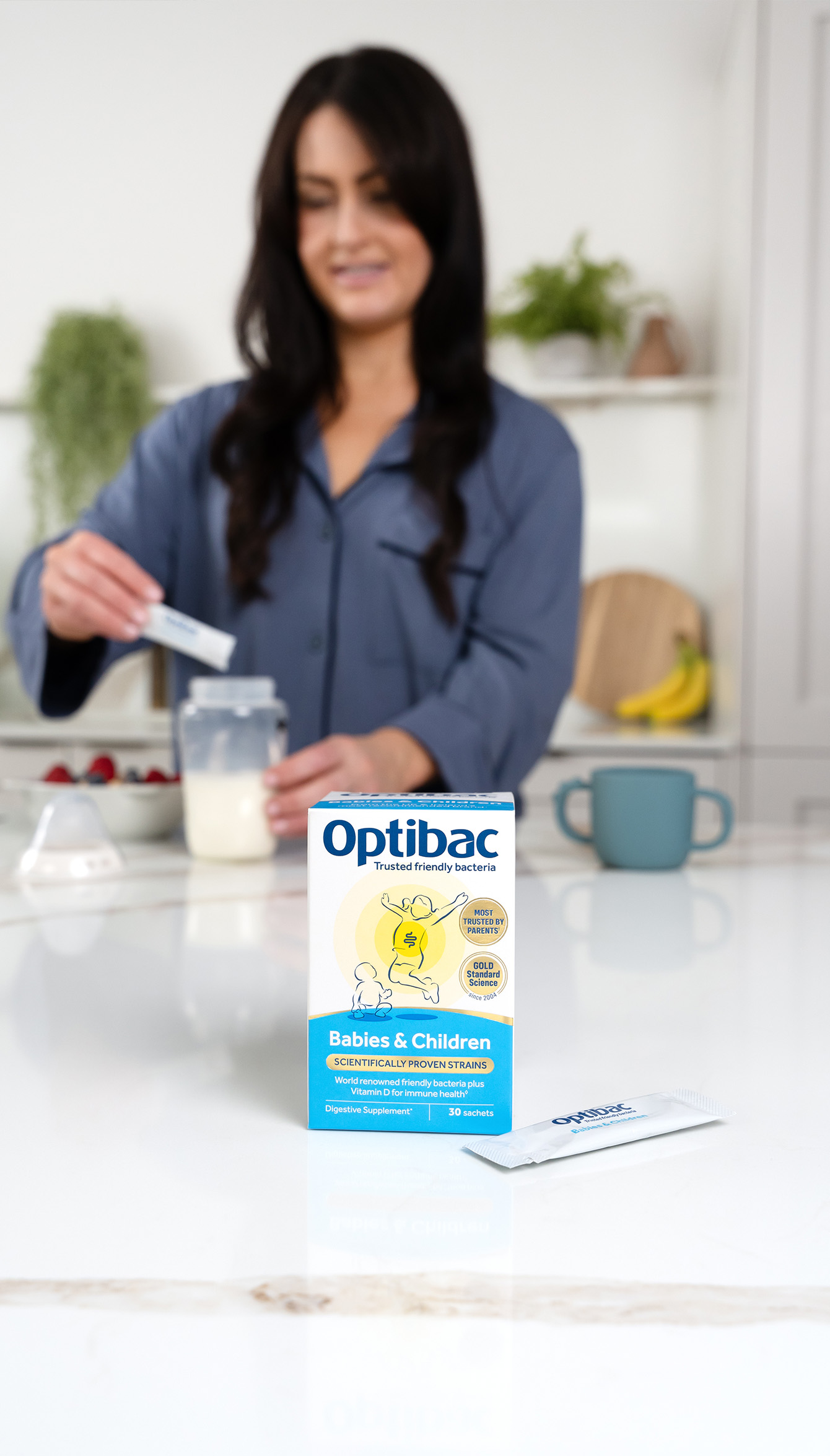 A woman in a kitchen prepares a baby bottle. In the foreground, Optibac probiotics for babies and children is displayed.