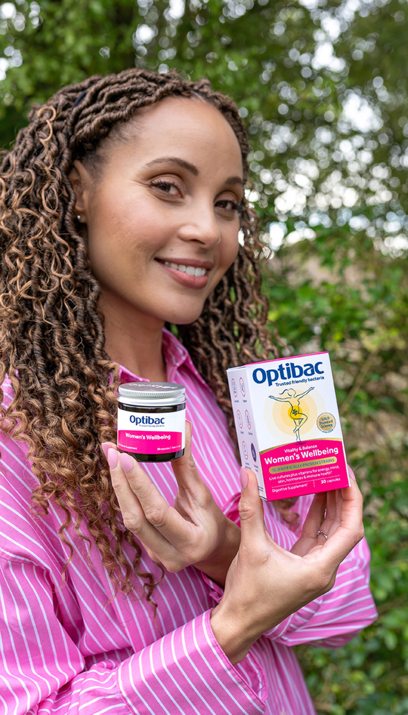 Woman in a pink shirt smiling and holding Optibac Women's Wellbeing outdoors, with greenery in the background.