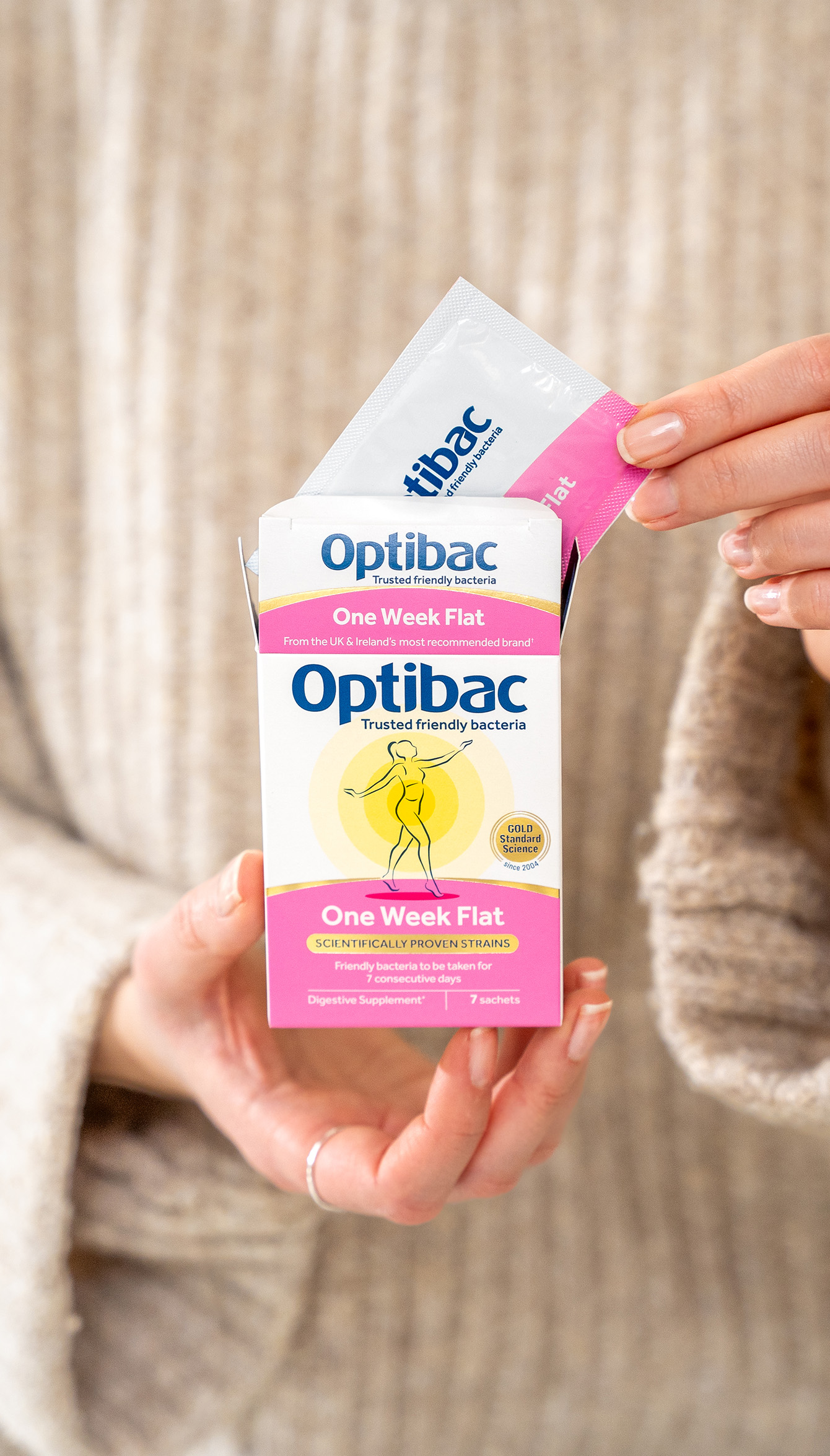 A person holding a box of Optibac One Week Flat probiotics, with a sachet being removed. The packaging features a yellow design and pink accents.