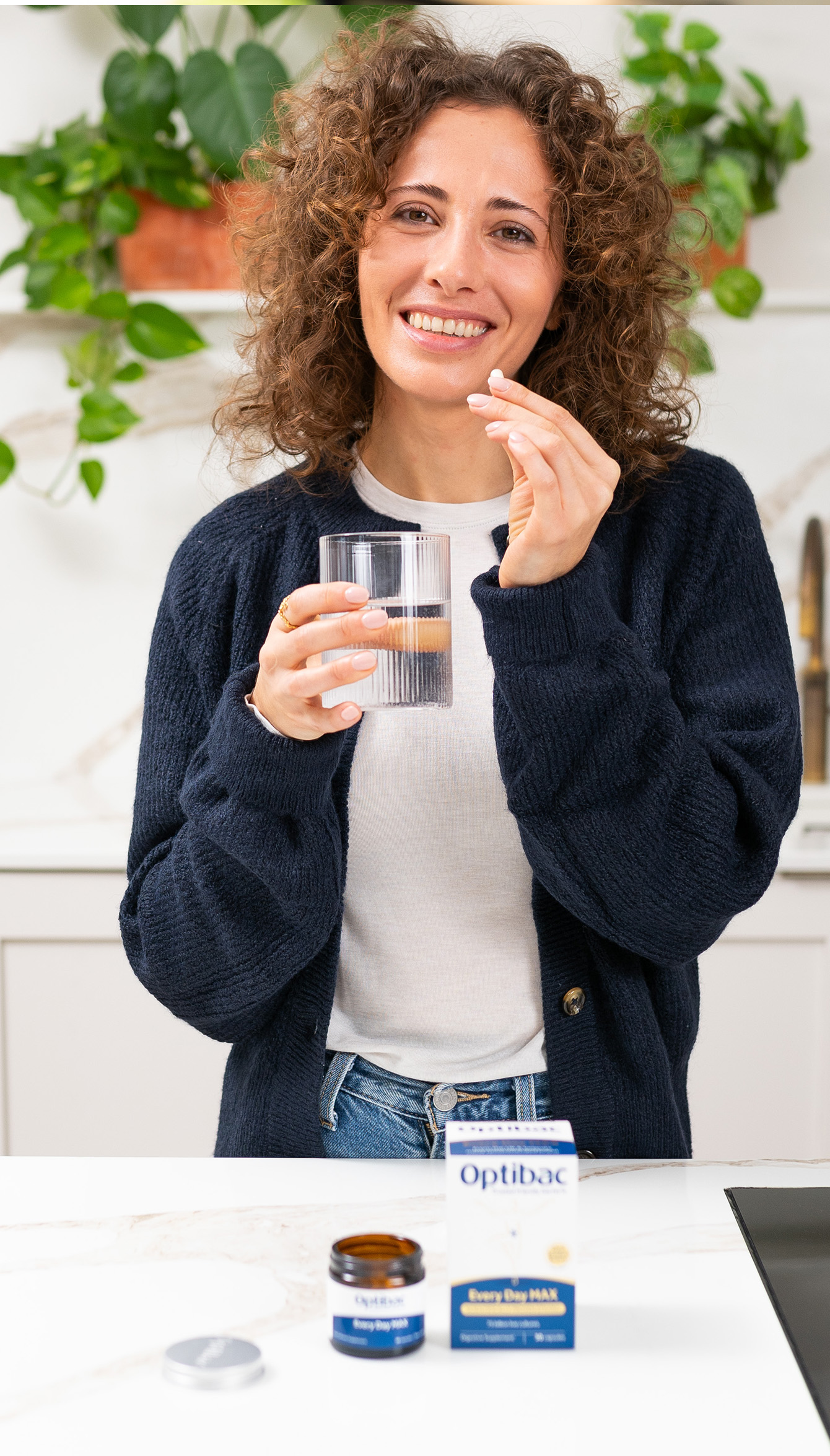 A person with curly hair smiles while holding a glass of water and a pill, with Optibac supplements on the counter in a kitchen setting.