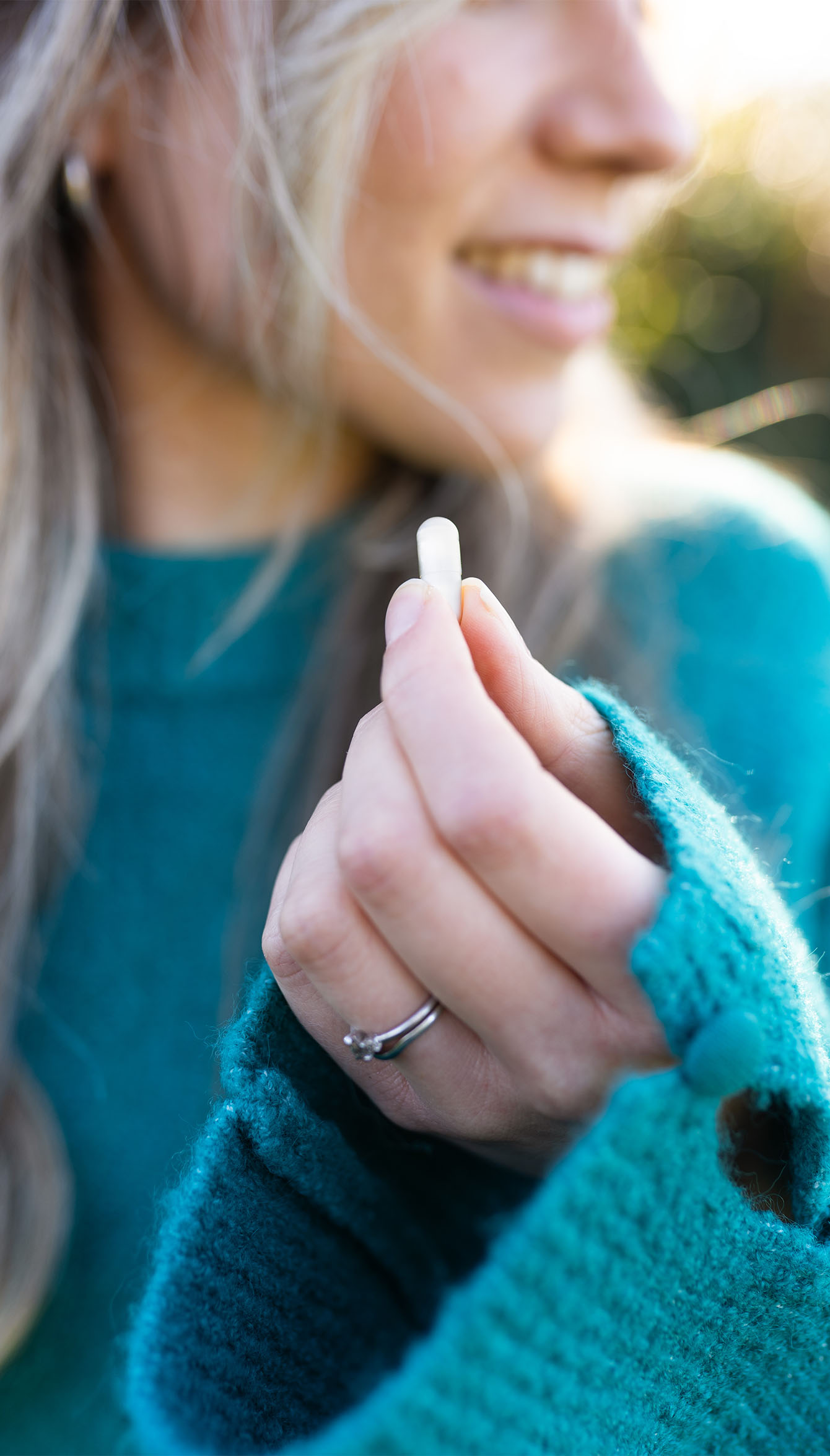 Woman in a teal sweater holding a small white pill between her fingers, smiling slightly, with sunlight in the background.