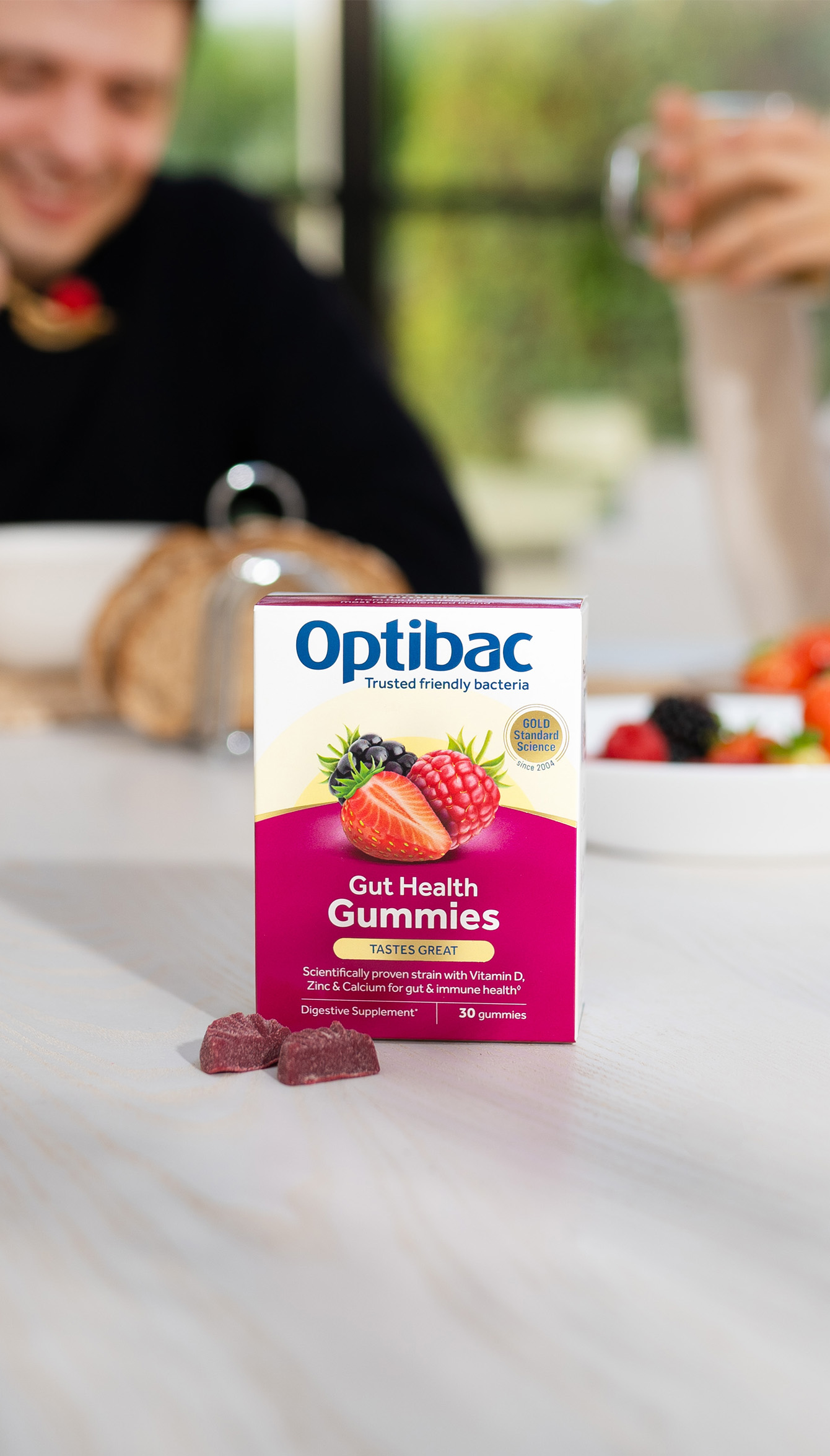 Box of Optibac Gut Health Gummies on a table with two gummies in front, blurred background with people and a bowl of fruit.