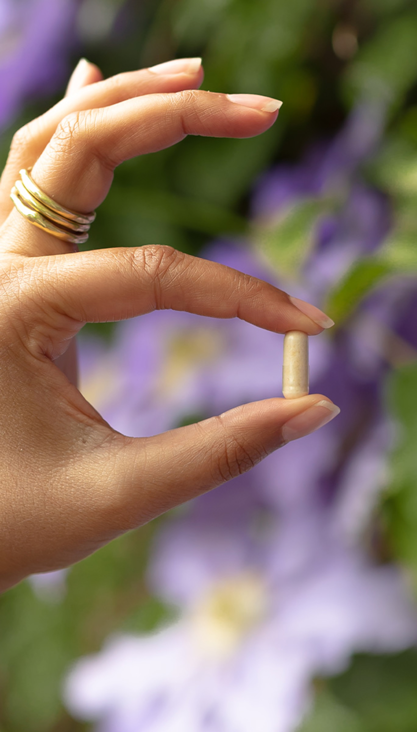 A hand with gold rings holds an Optibac capsule against a blurred background of purple flowers.