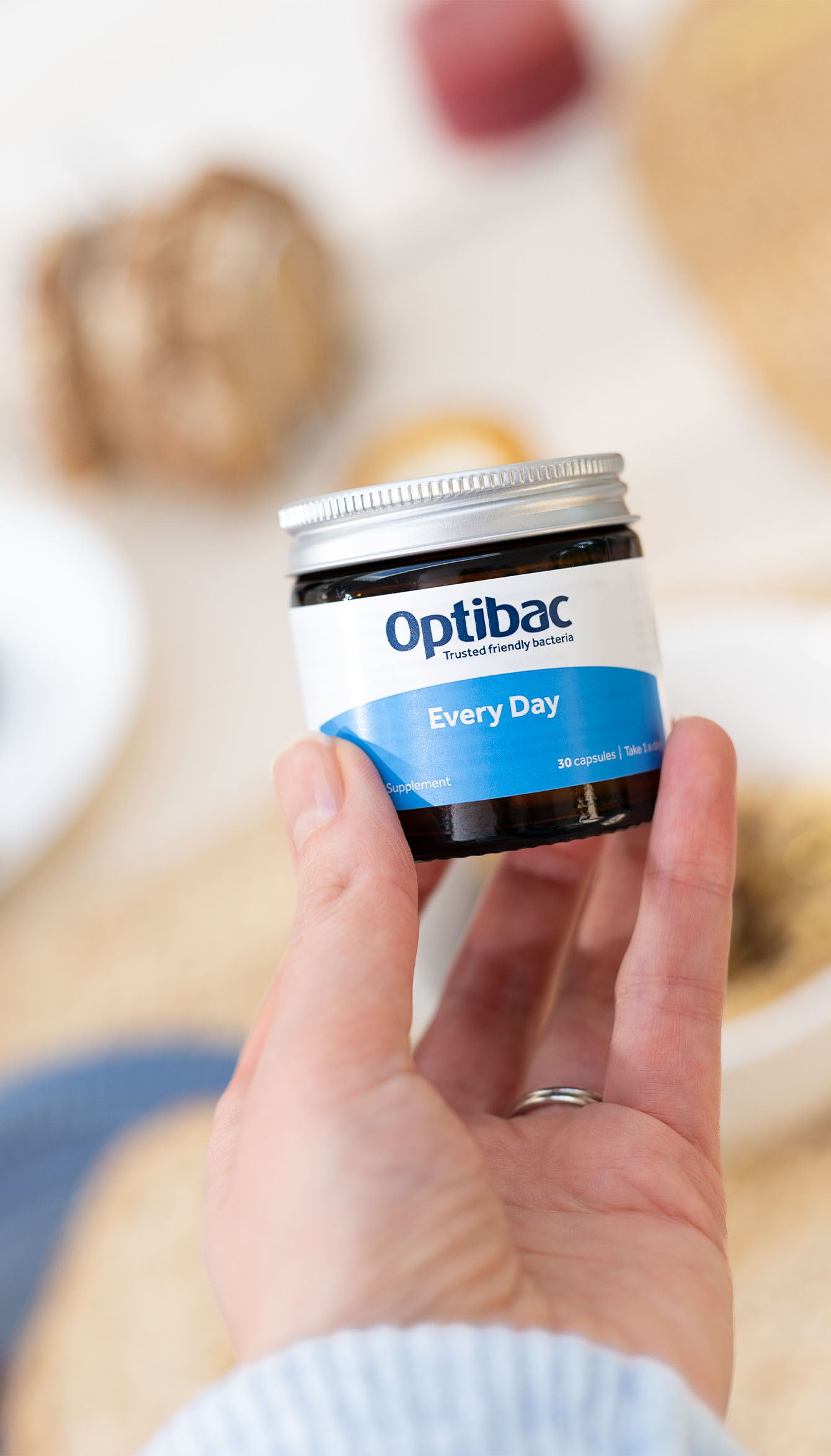 Hand holding a jar of Optibac Every Day capsules, with a blurred background of household items.