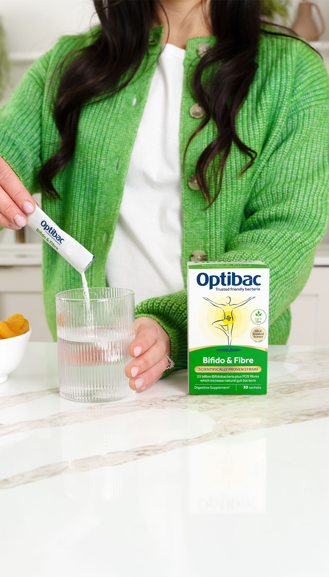 Woman in a green sweater pours Optibac Bifido & Fibre sachet into a glass of water on a kitchen counter.