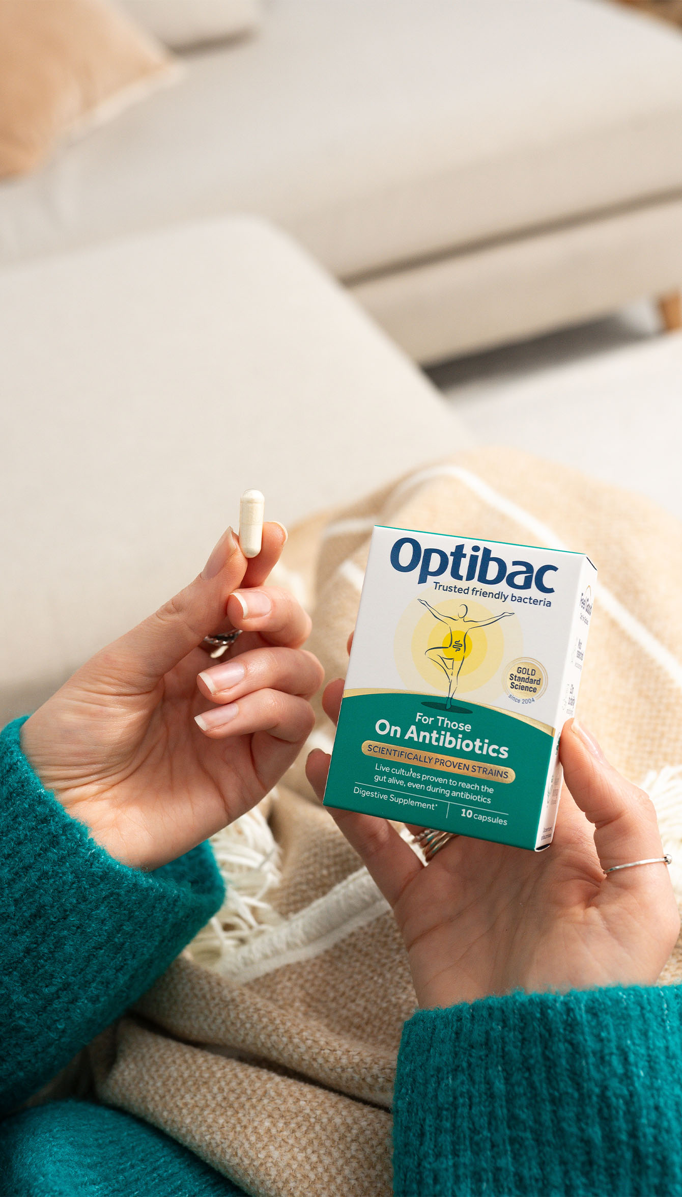 Person holding a capsule and a box of Optibac probiotics, sitting on a beige couch with a blanket.