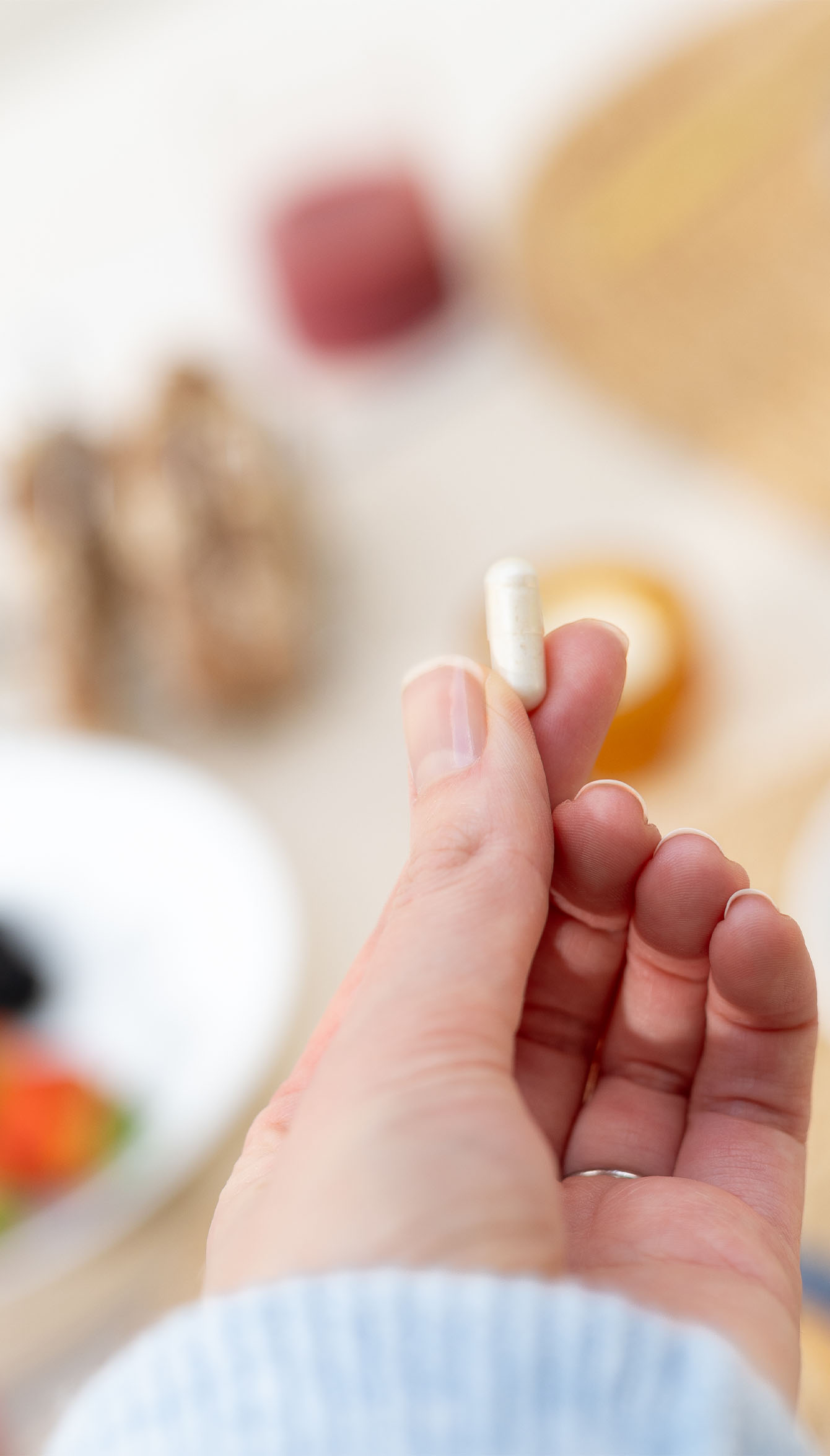 A hand holding a white capsule pill with a blurred background featuring a plate of food and various items.