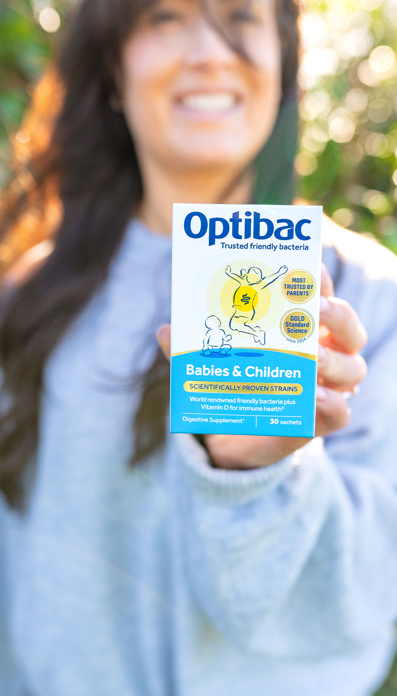 Person holding a box of Optibac Babies & Children probiotics outdoors, with a blurred background of greenery.