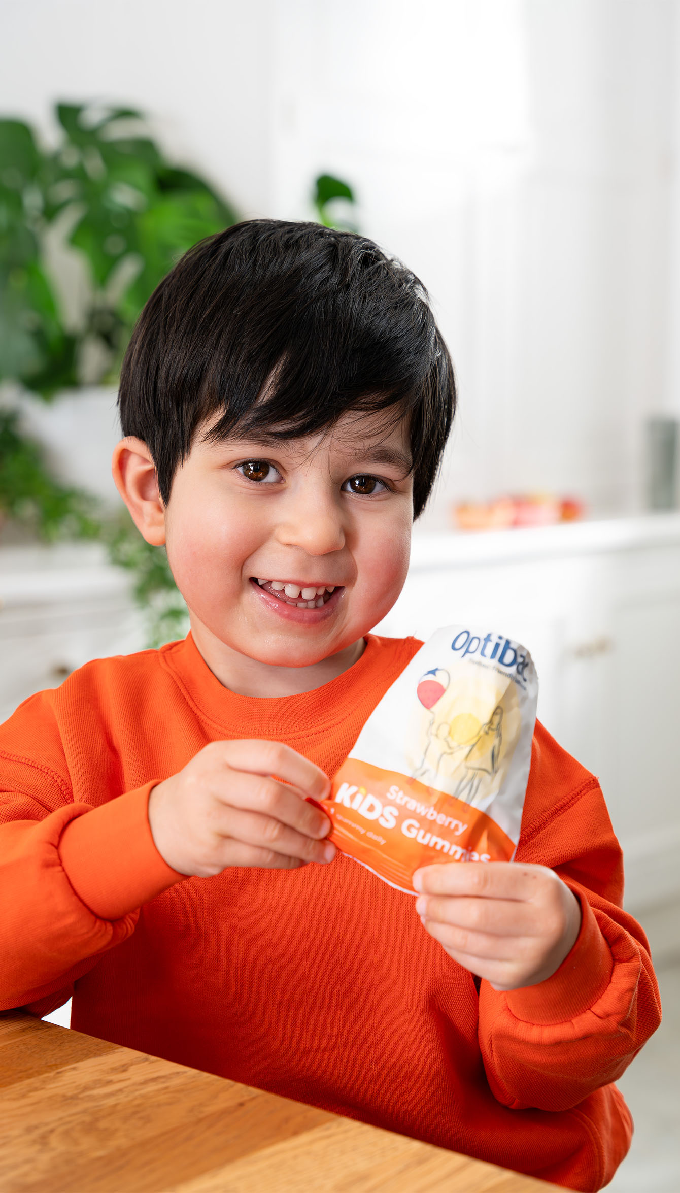 Smiling child in an orange shirt holding a packet of strawberry-flavored kids' gummies in a bright kitchen.