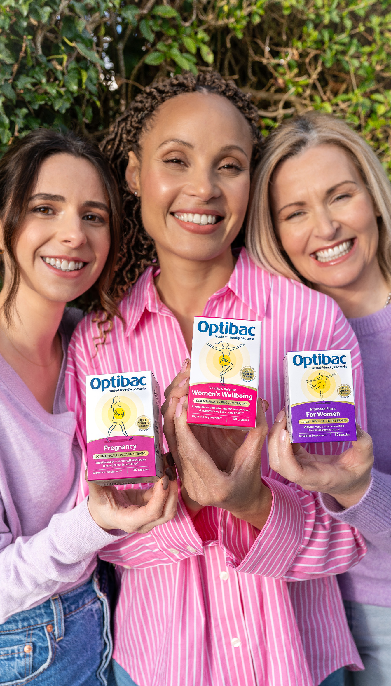Three smiling women hold Optibac probiotic boxes outdoors, showcasing products for pregnancy, women's wellbeing, and general women's health.