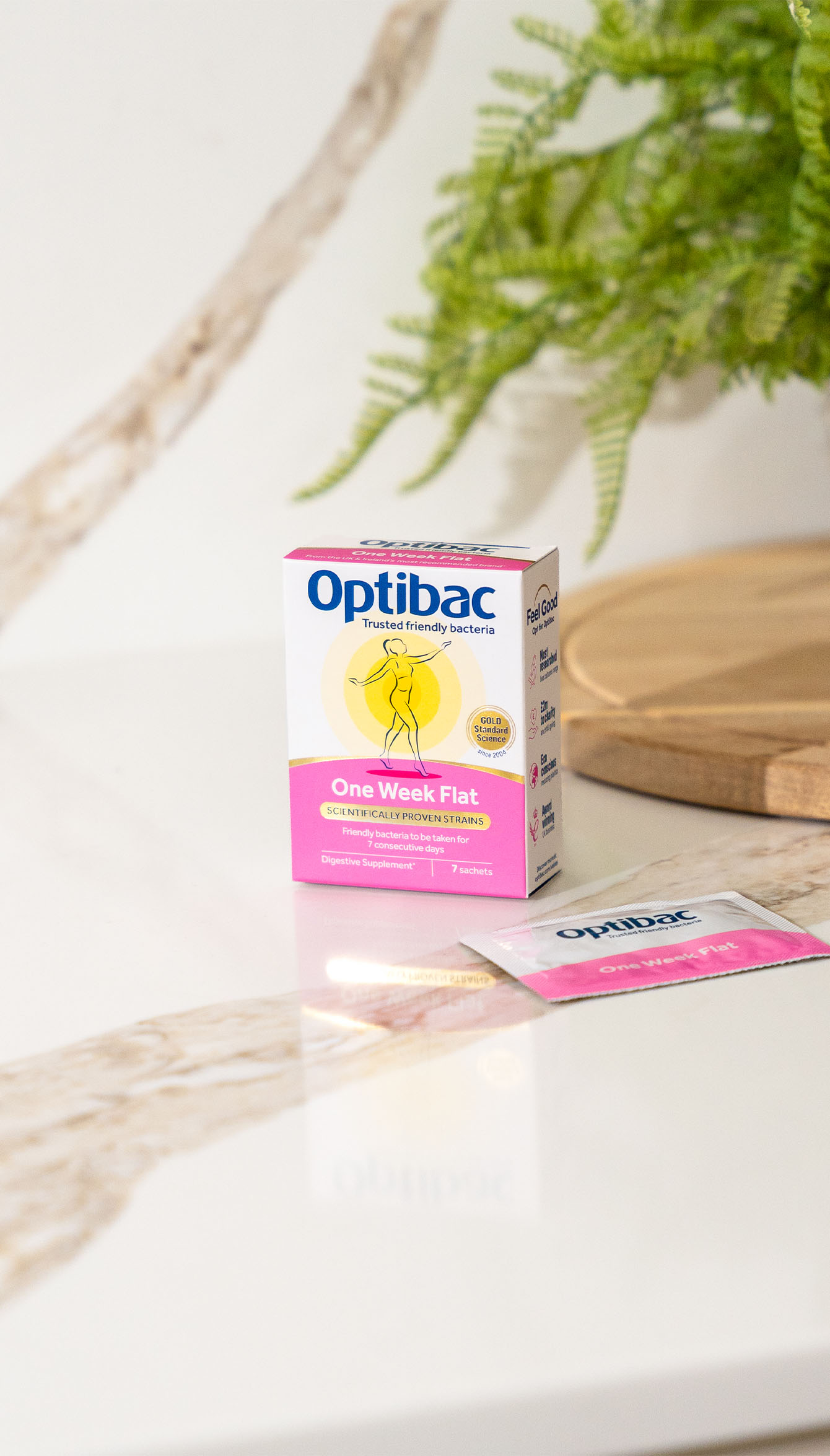 Box of Optibac One Week Flat probiotics and a sachet on a marble surface, with a wooden tray and green plant in the background.
