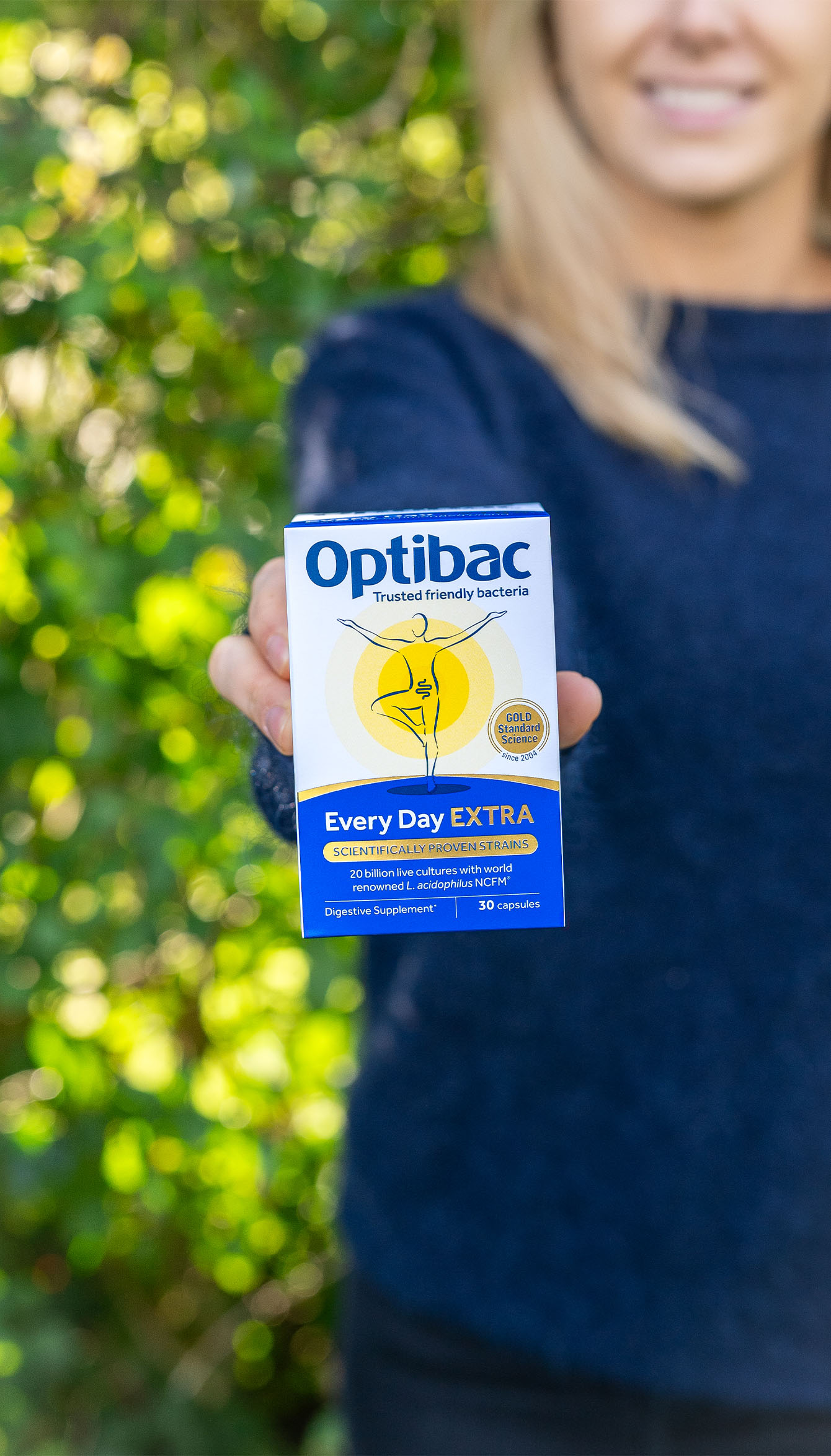Person holding a box of Optibac Every Day EXTRA probiotics against a green leafy background.