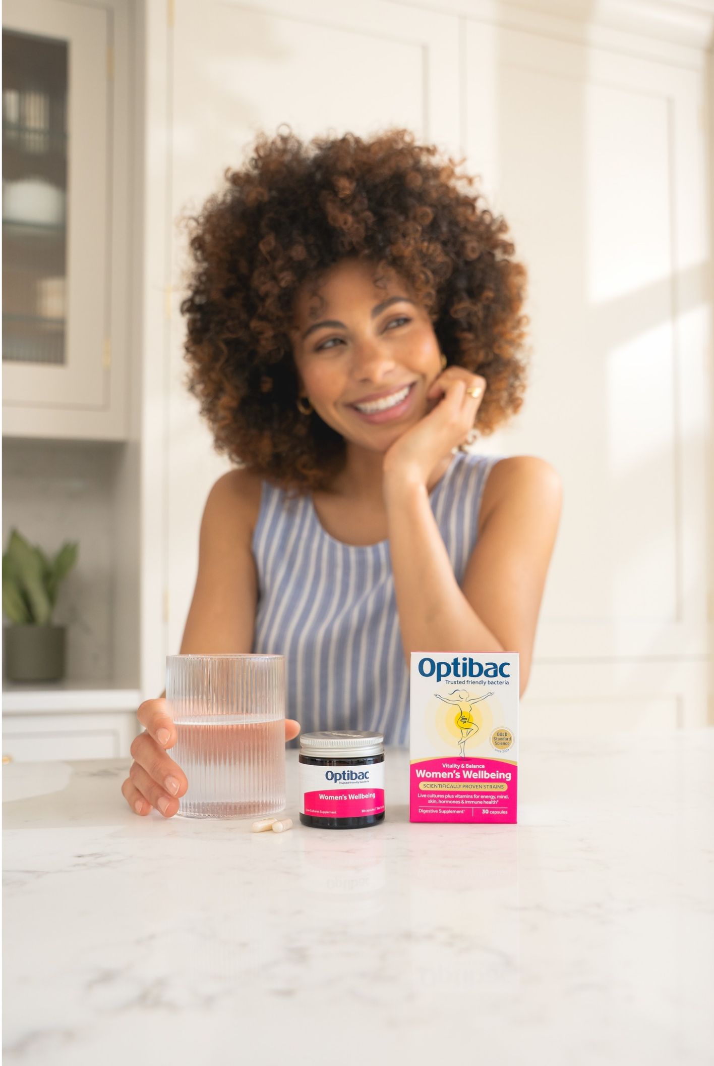 Woman smiling at a kitchen counter with a glass of water, Optibac probiotic capsules, and packaging labeled "Women's Wellbeing" in front of her.