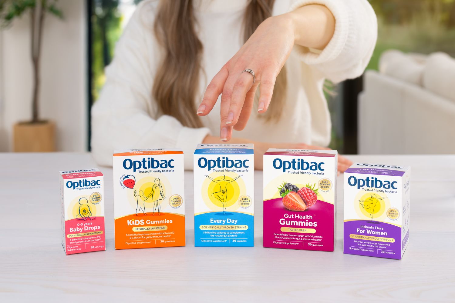 A person selects one of five Optibac probiotic product boxes on a table, featuring varieties for babies, kids, daily use, gut health, and women.