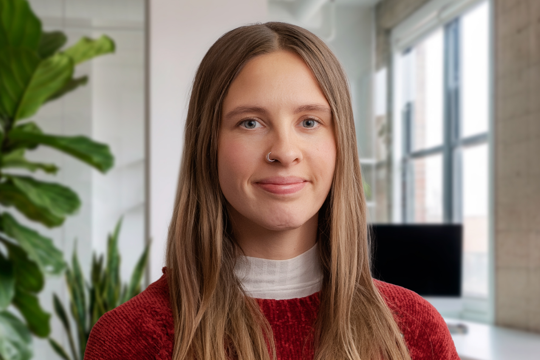 A woman with long brown hair and a nose ring smiling, wearing a red sweater in a modern office with plants in the background.