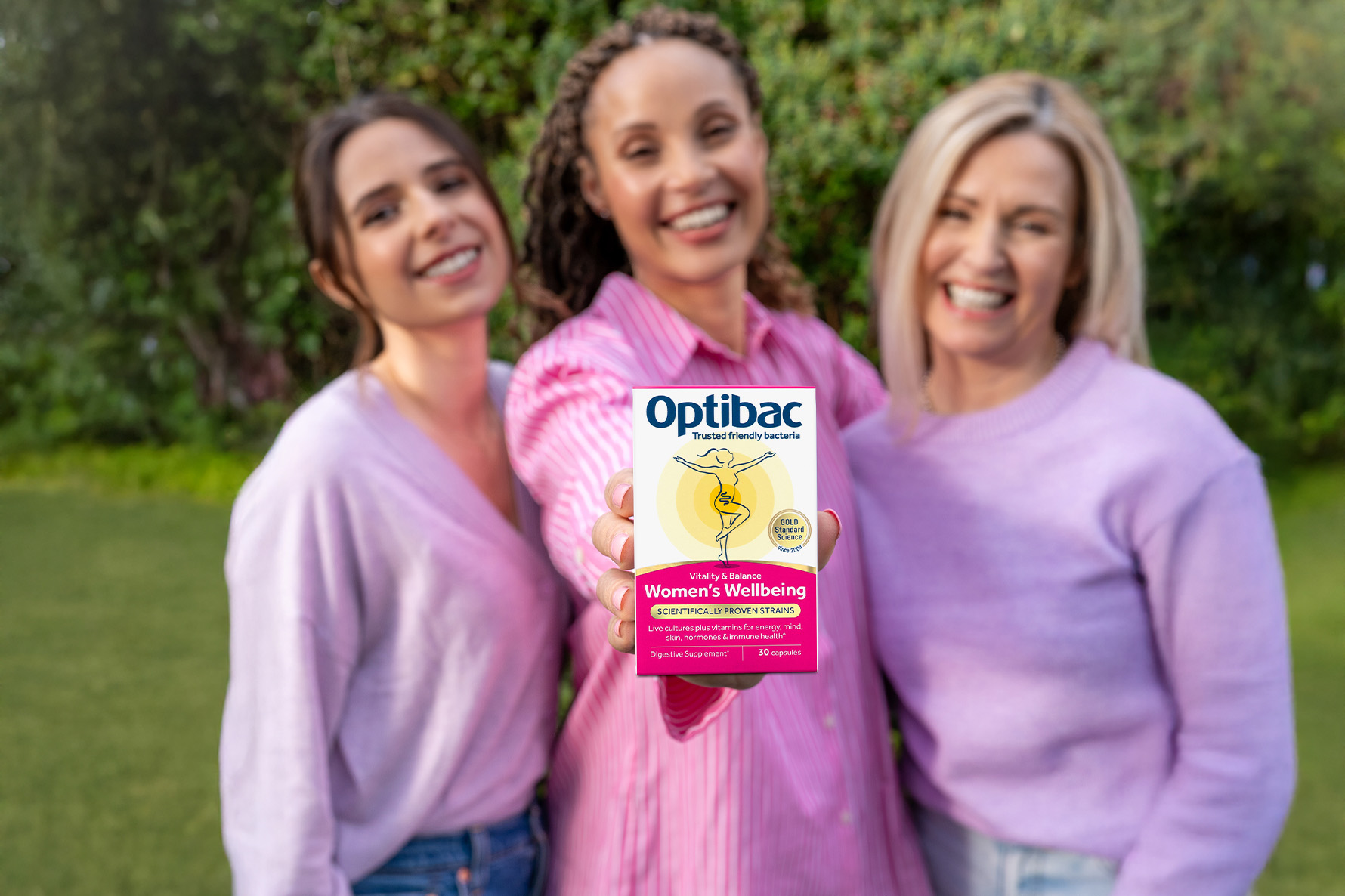 Three women smiling outdoors, one holding a box of Optibac Women's Wellbeing supplements, with greenery in the background.
