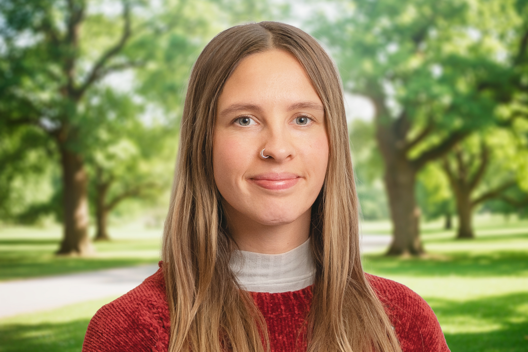 Smiling person with long hair and a nose ring, wearing a red sweater, standing in a sunlit park with green trees in the background.