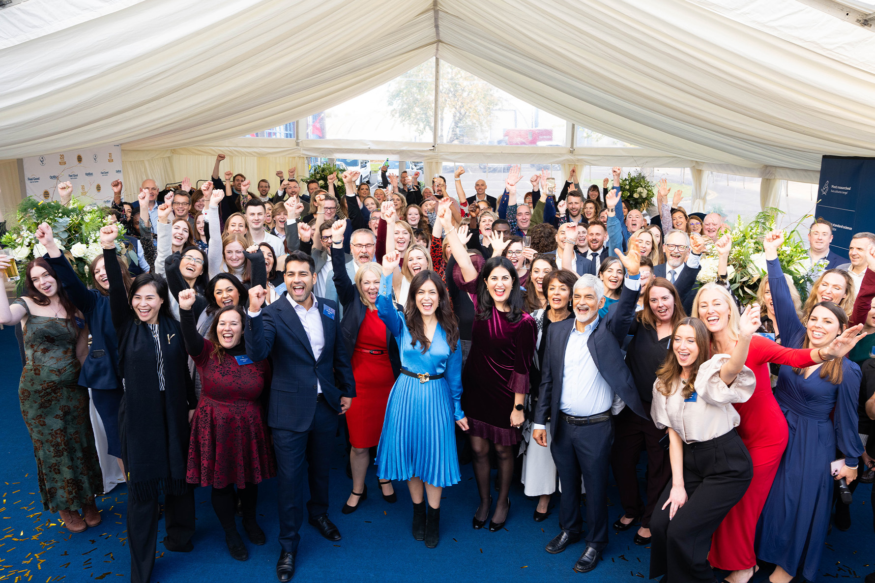 A large group of people in a celebratory pose under a white tent, smiling and raising their hands, with floral arrangements in the background.