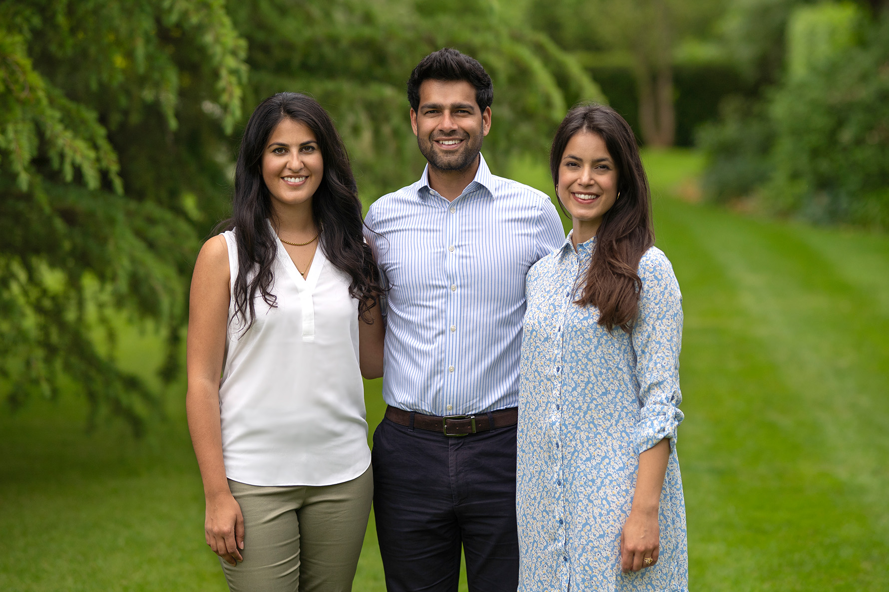 Three people standing on a grassy path with trees in the background, smiling at the camera.