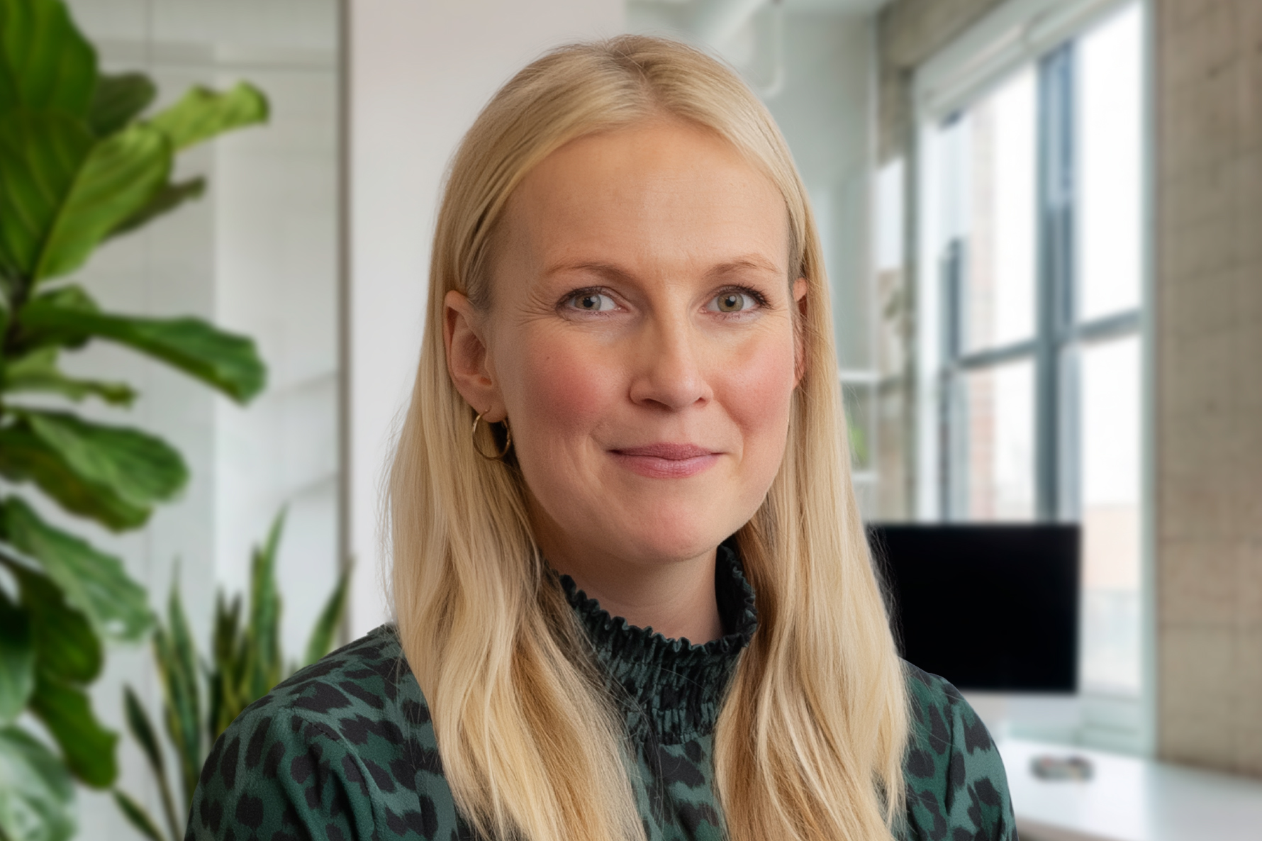A woman with long blonde hair and wearing a green patterned top smiles slightly in a bright office with plants and large windows.