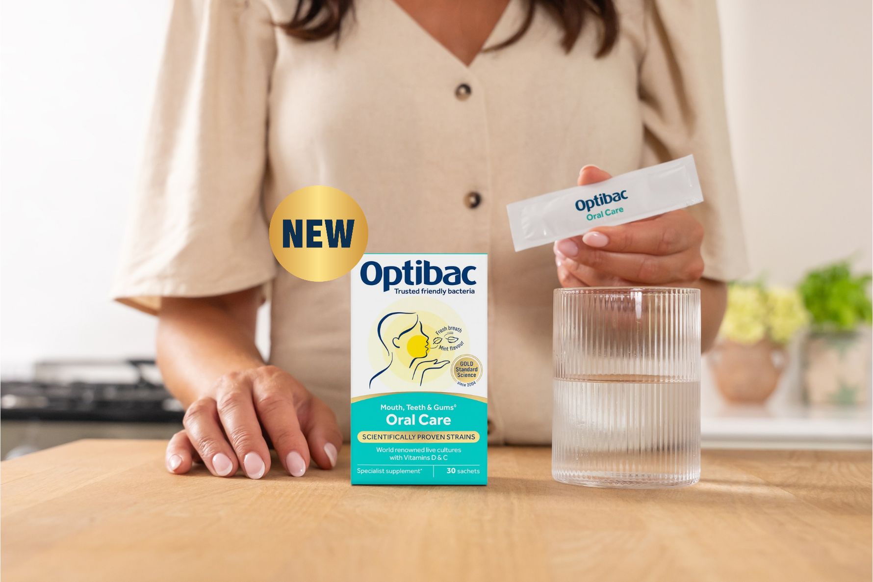 A person holding an Optibac Oral Care sachet next to a glass of water, with a box labeled "Optibac Oral Care" on a table.