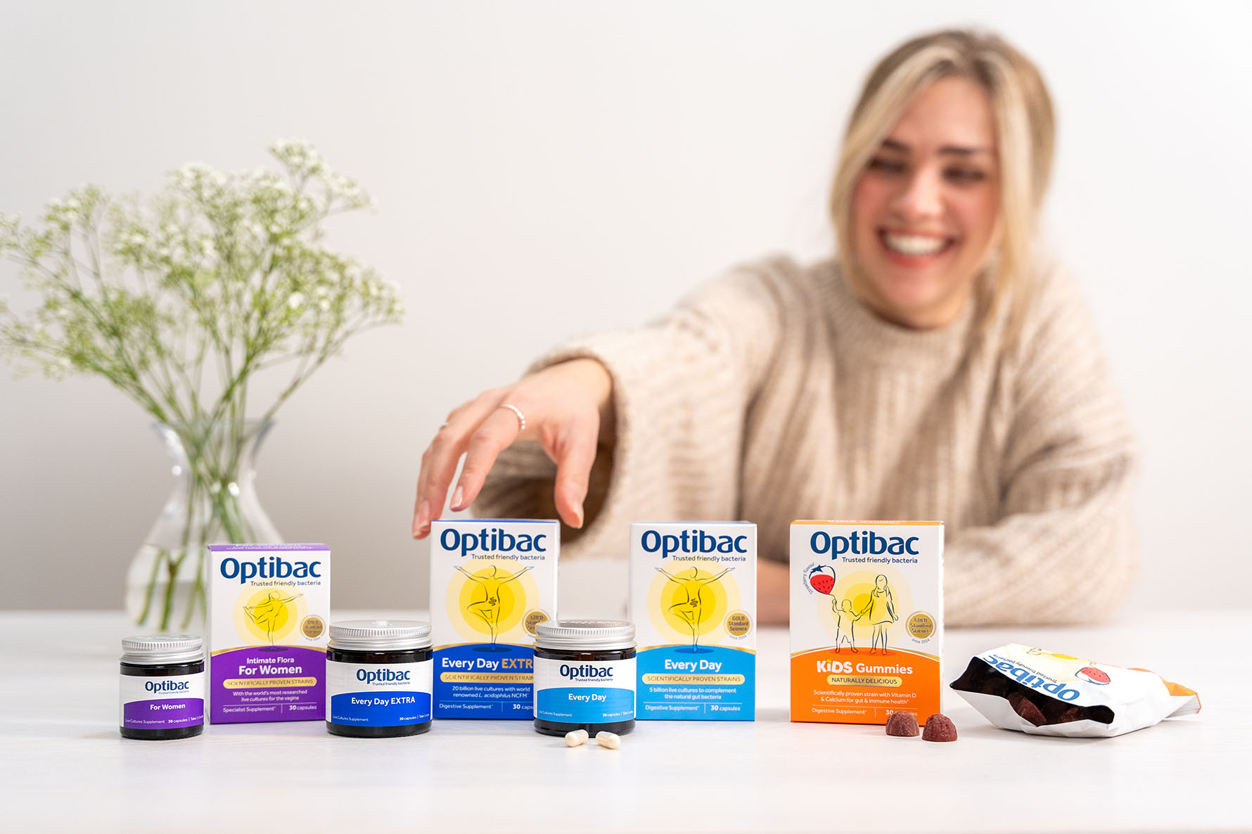 Smiling woman reaching for Optibac probiotic products on a table, with a vase of flowers in the background.