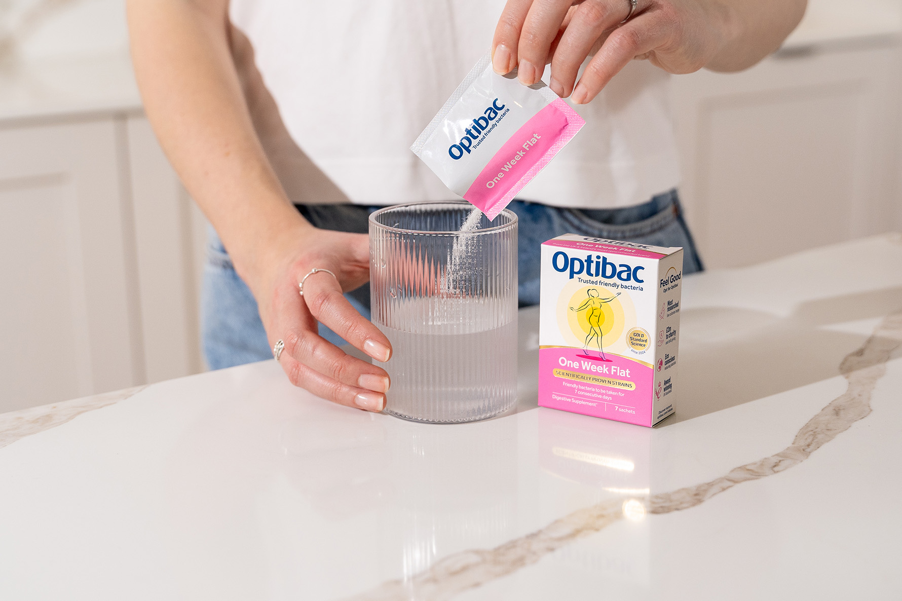 Person pouring Optibac probiotic powder into a glass of water on a marble countertop, with the product box nearby.