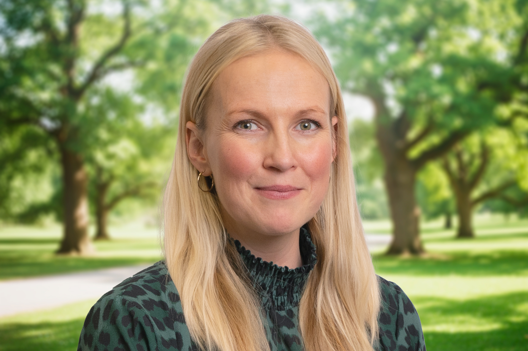 Woman with long blonde hair and hoop earrings smiling, standing in a park with green trees in the background.