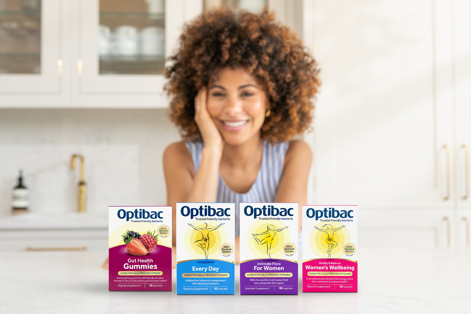 Smiling woman in a kitchen with four boxes of Optibac supplements displayed in front of her.