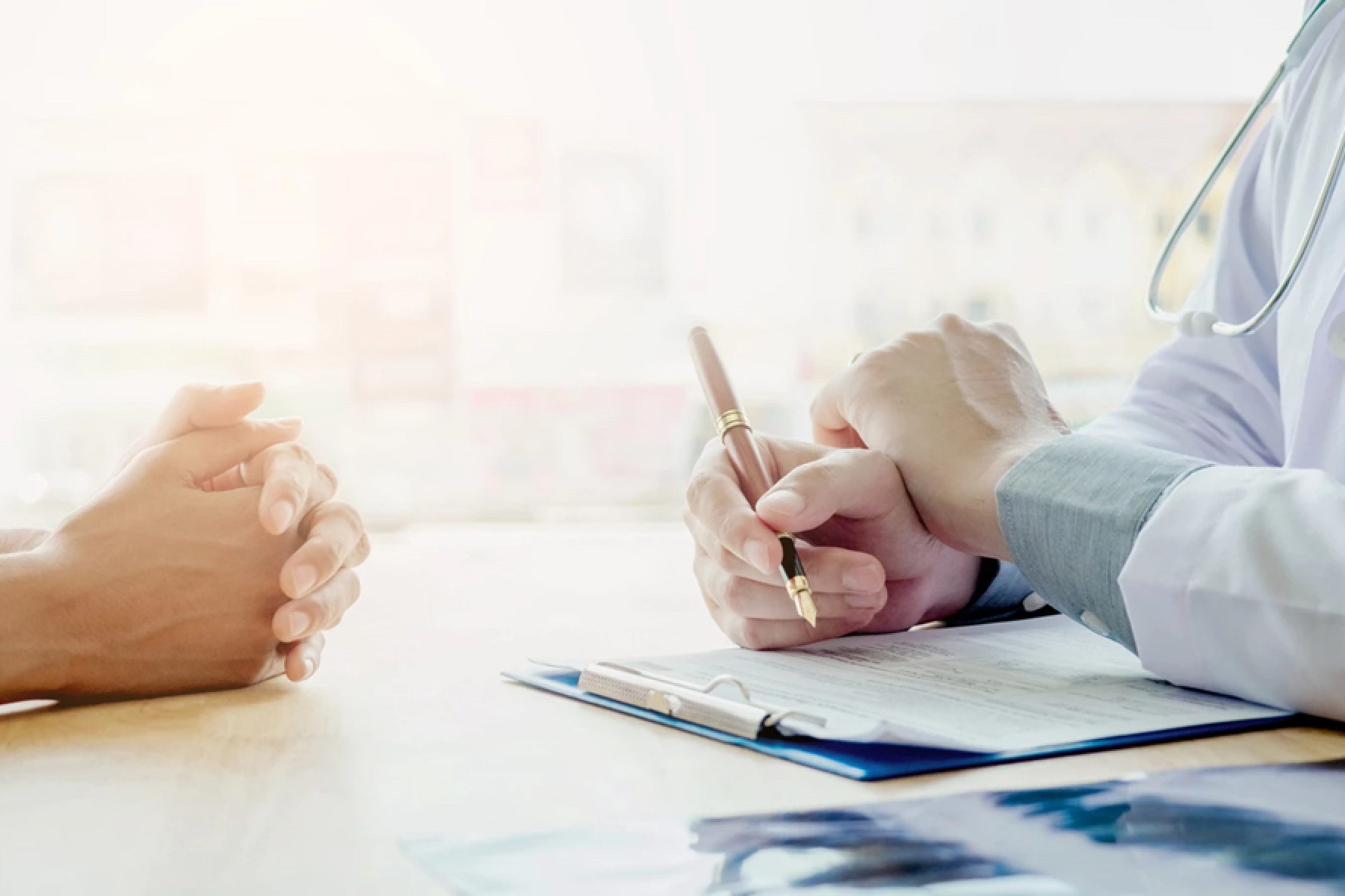 Doctor discussing with a patient, holding a pen over a clipboard. Both have hands resting on a table in a well-lit room.
