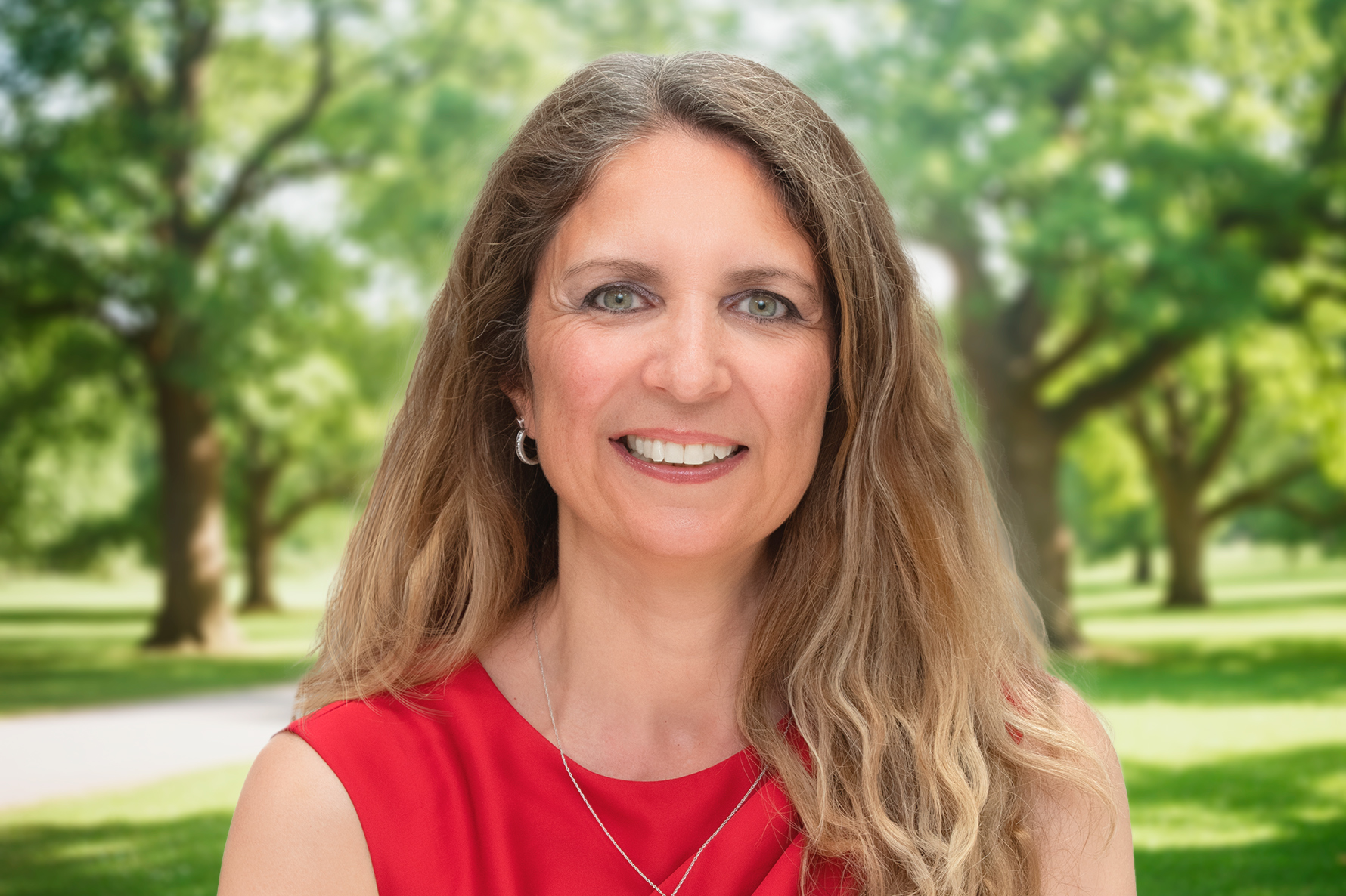 Smiling woman with long hair in a red top stands in a sunlit park with green trees in the background.