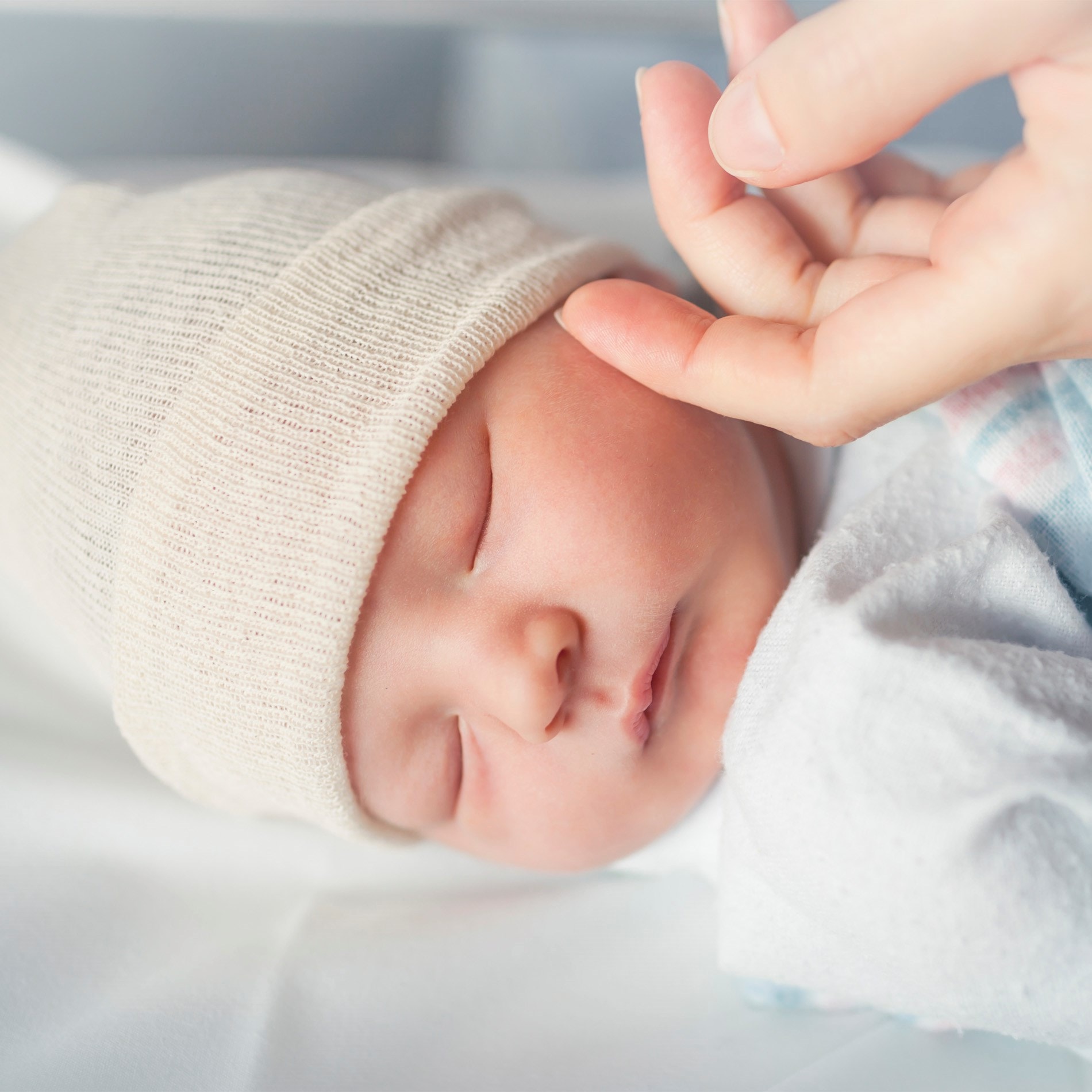 Sleeping newborn in a beige hat, gently touched by an adult hand, wrapped in a soft blanket on a white surface.