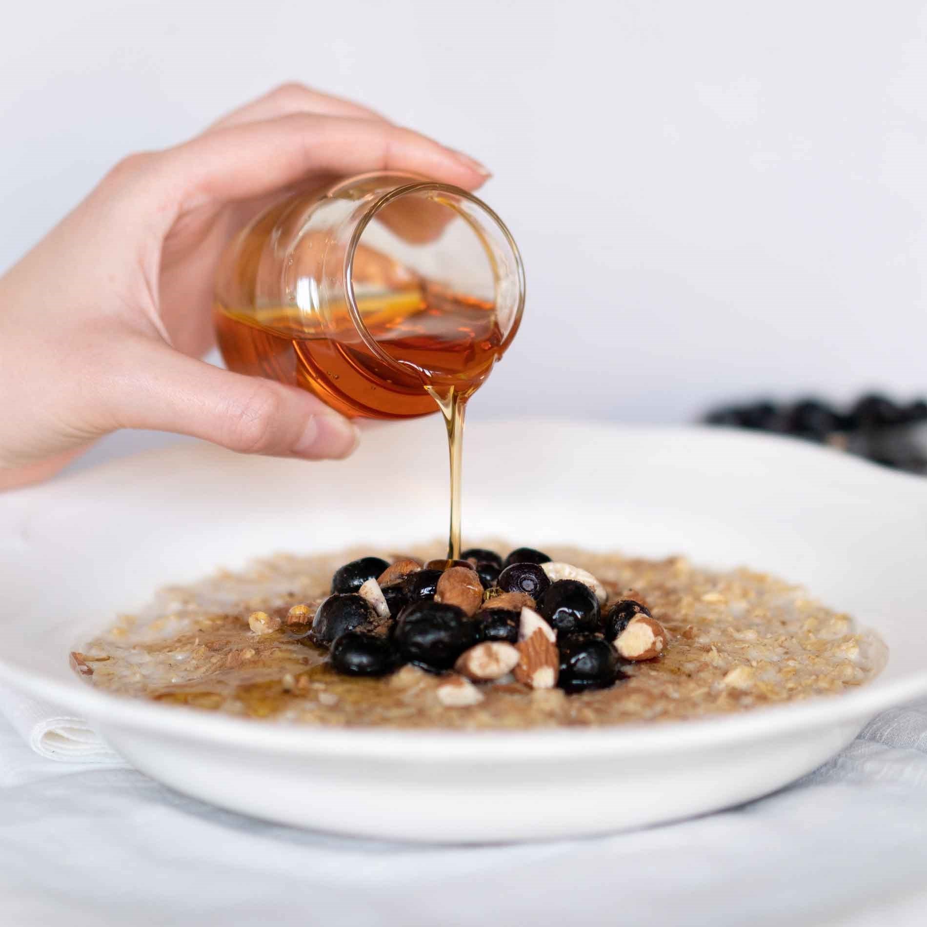 Hand pouring honey over a bowl of oatmeal topped with blueberries and almonds on a white background.