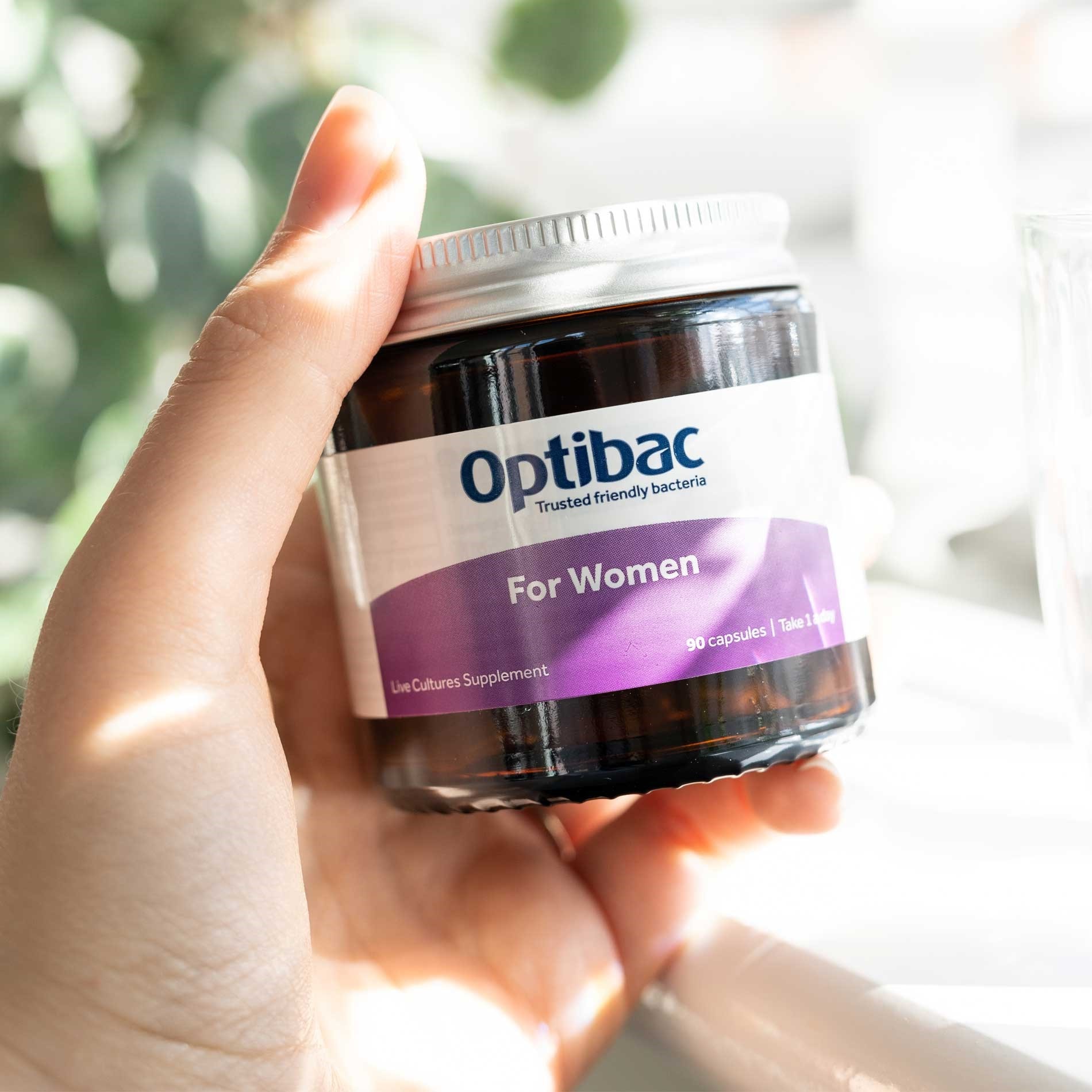 Hand holding a jar of Optibac For Women live cultures supplement with a purple label, against a blurred background of plants.